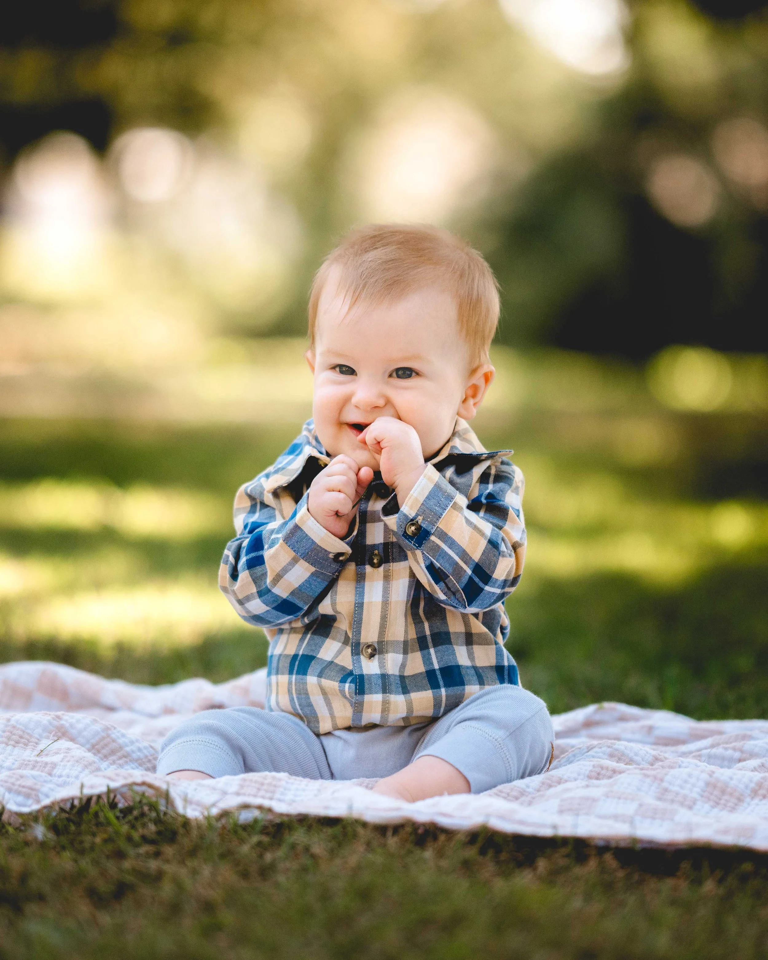 A baby sitting on a blanket outdoors, wearing a plaid shirt, smiling, and holding his hand near his mouth.