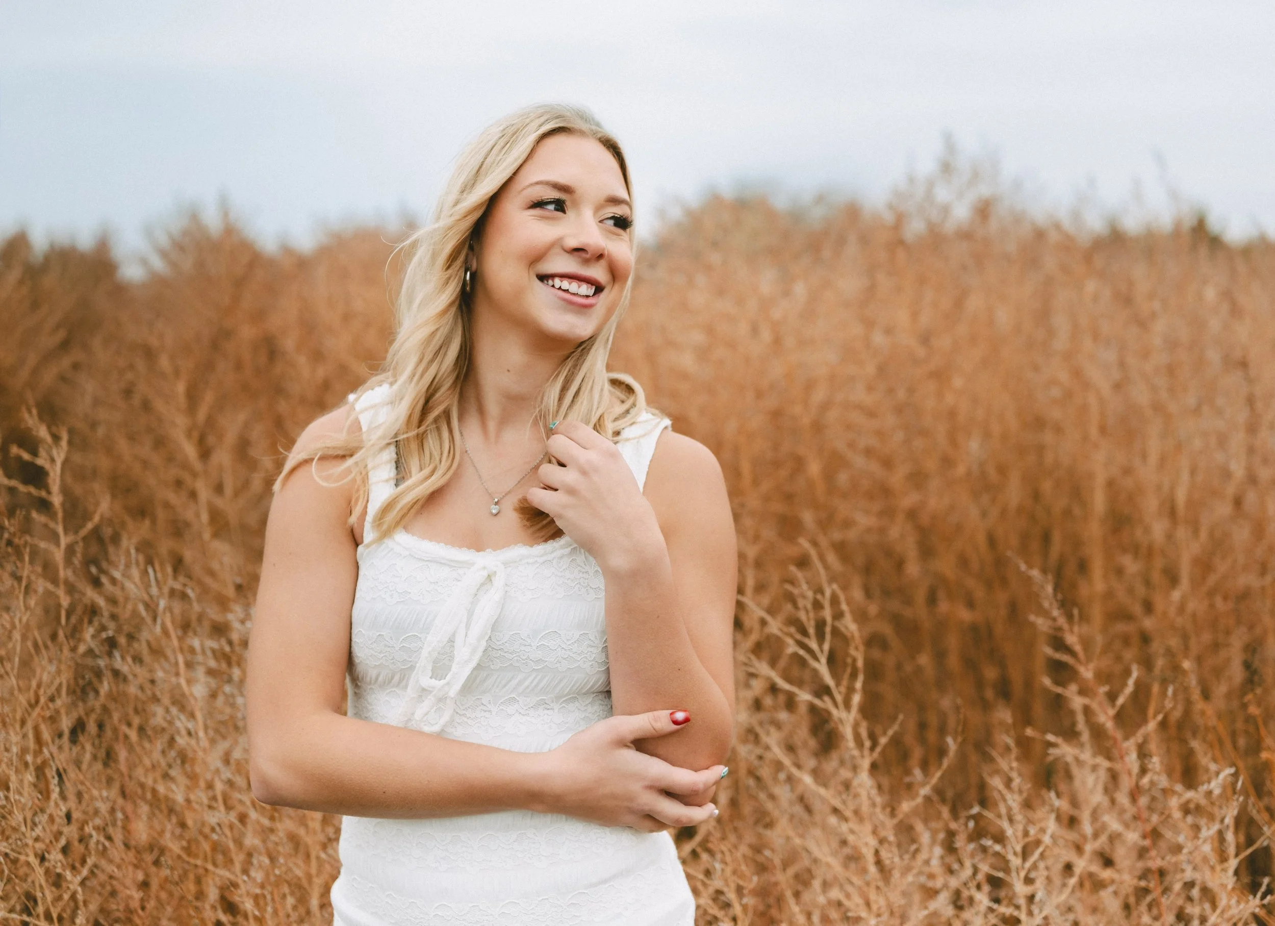 A young woman with blonde hair standing in a field of dry, brown plants, smiling and looking to her left.