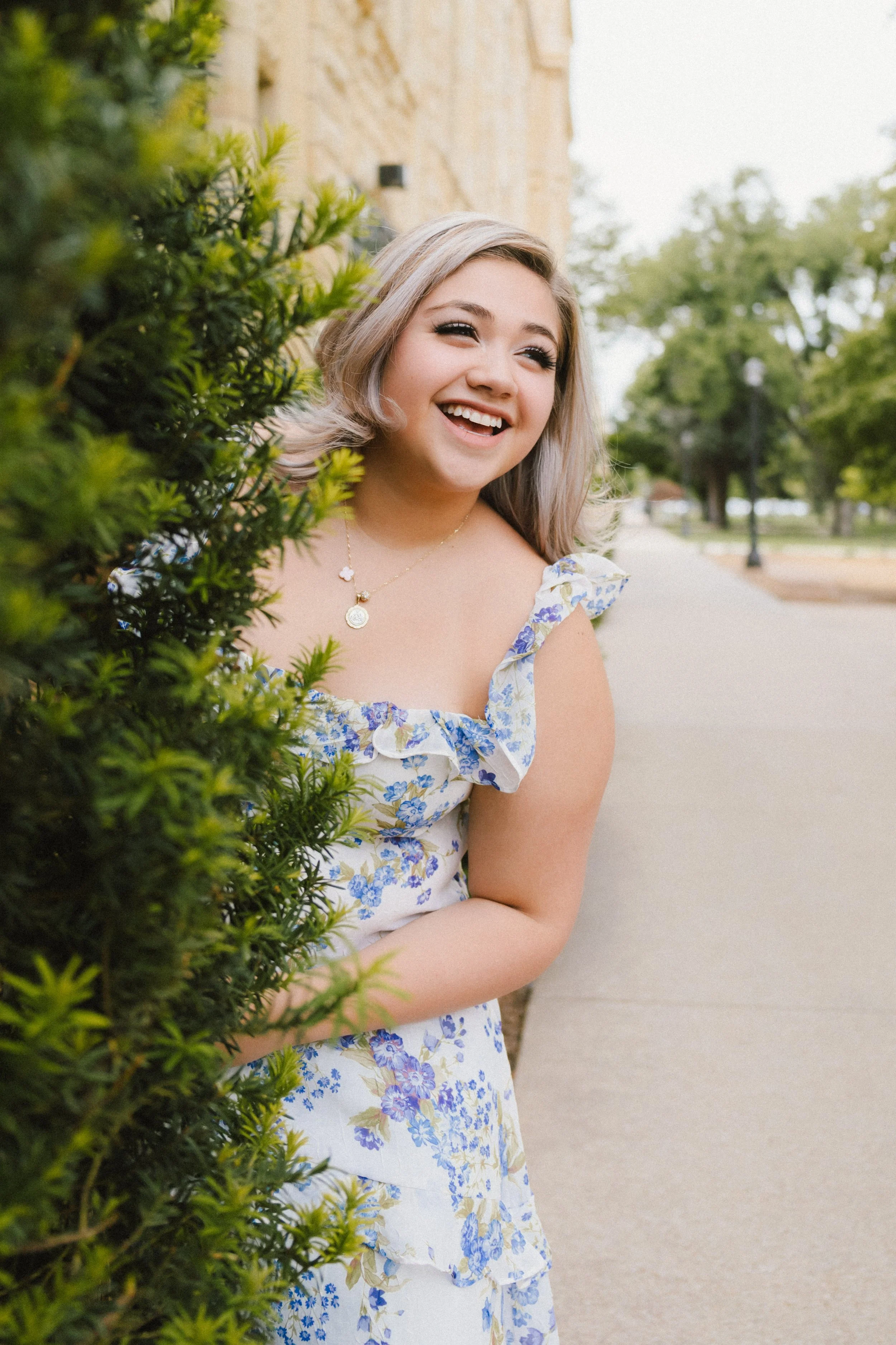 A young woman with blonde hair and a floral dress smiling and looking to her right, partially hidden behind green bushes on the left side of the image, in an outdoor park with trees and a pathway in the background.