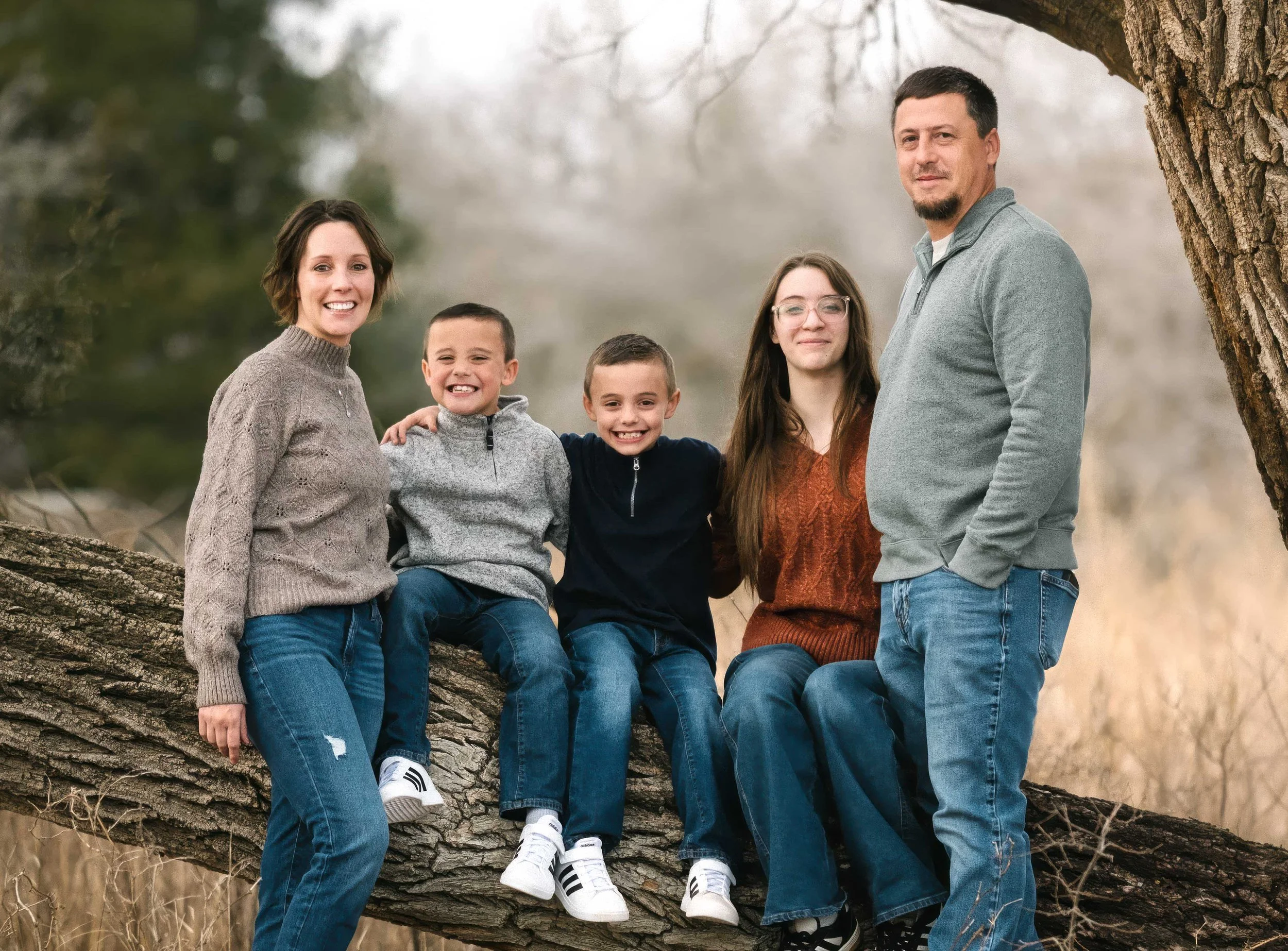 Family of five sitting on a large tree branch outdoors, smiling at the camera, with a blurred natural background.