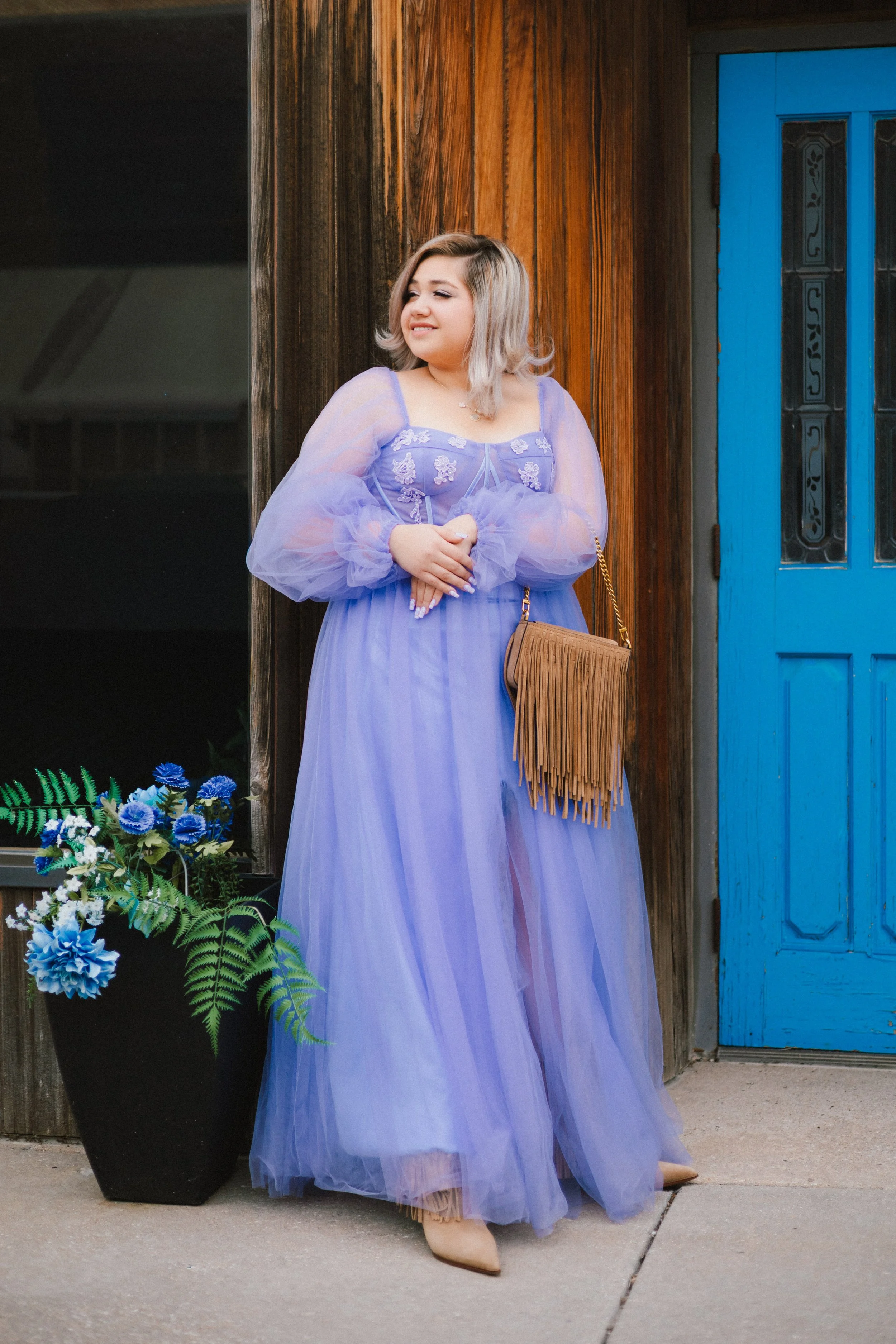 A woman with shoulder-length blonde hair standing next to a large black flowerpot filled with blue and white flowers, wearing a long lavender dress with sheer sleeves and beige high heels, in front of a wooden wall and a blue door.