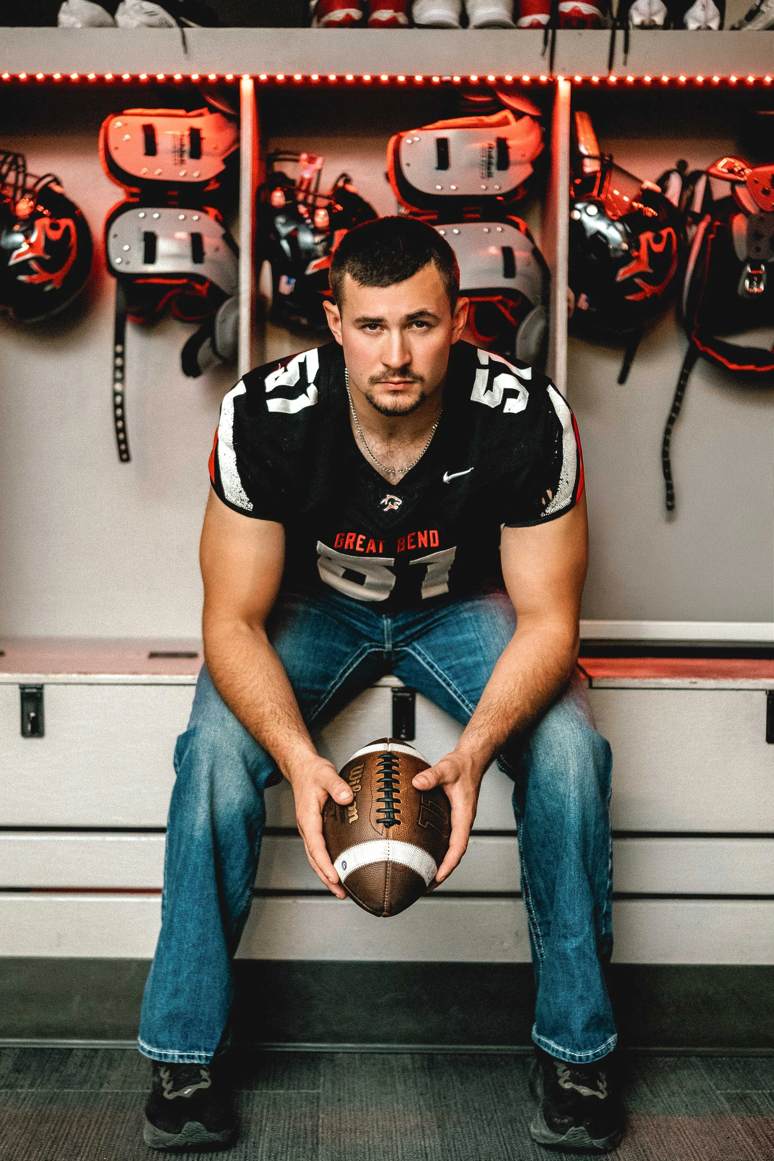A young man in a football uniform sitting on a bench in a locker room, holding a football, with football helmets hanging on the wall behind him.