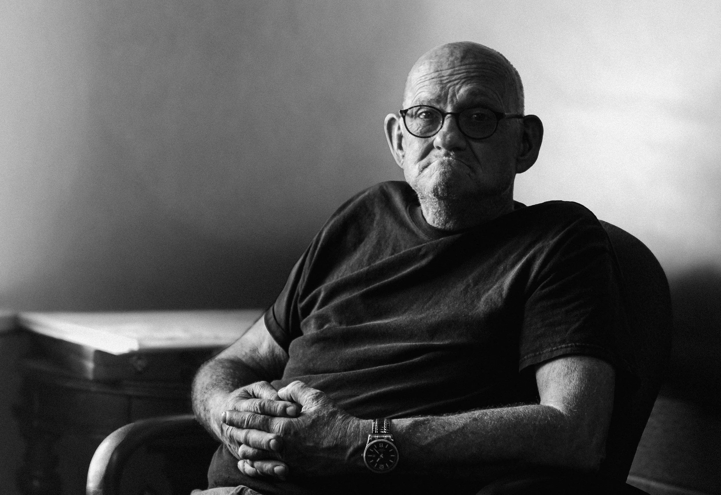 Black and white photo of an older man with glasses, short hair, and a goatee, sitting with his hands clasped on his lap, wearing a watch and casual T-shirt, looking directly at the camera against a plain background.