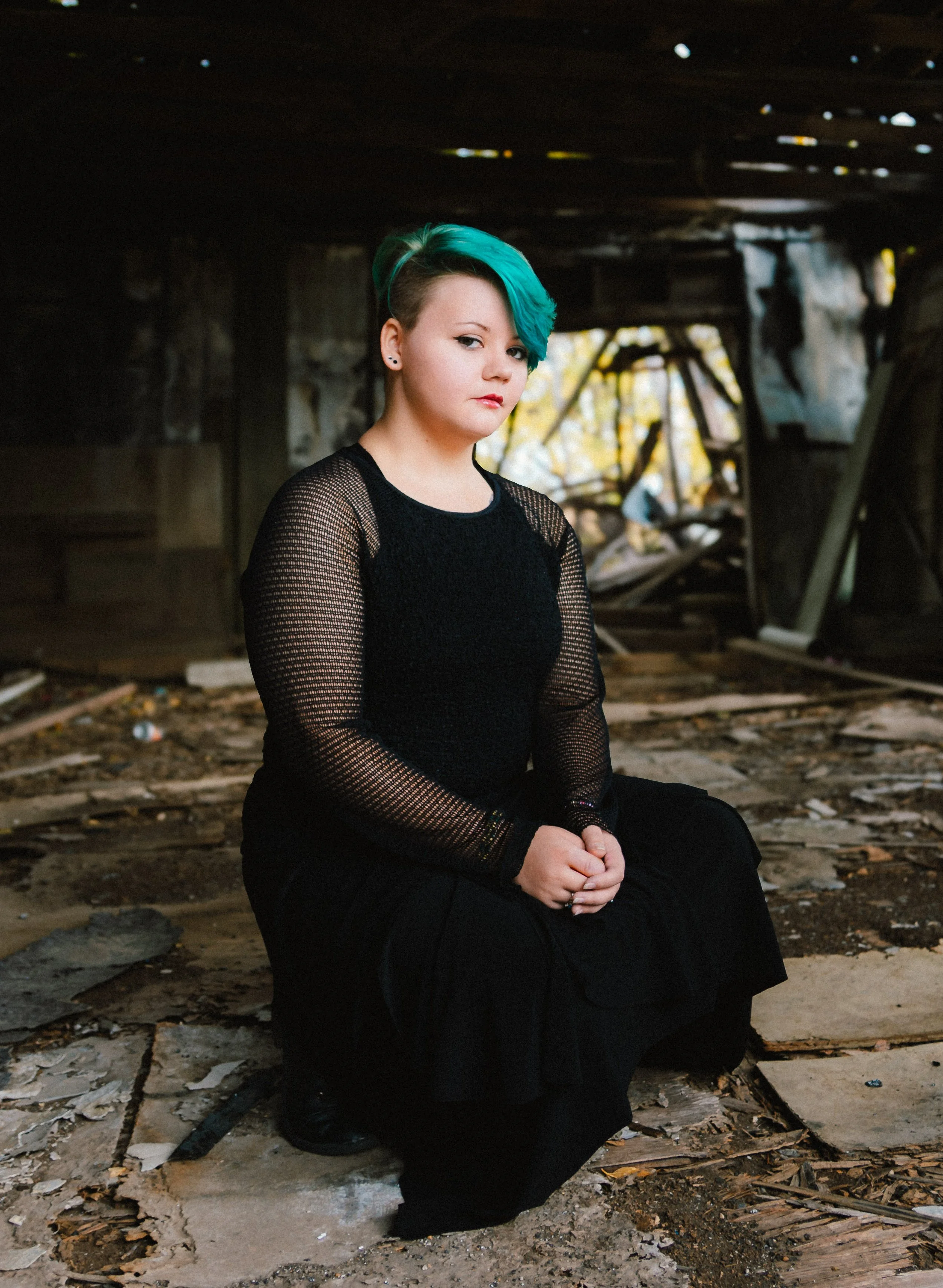 A woman with teal hair and a black mesh top squats on the ground inside a dilapidated building with broken wood and debris.
