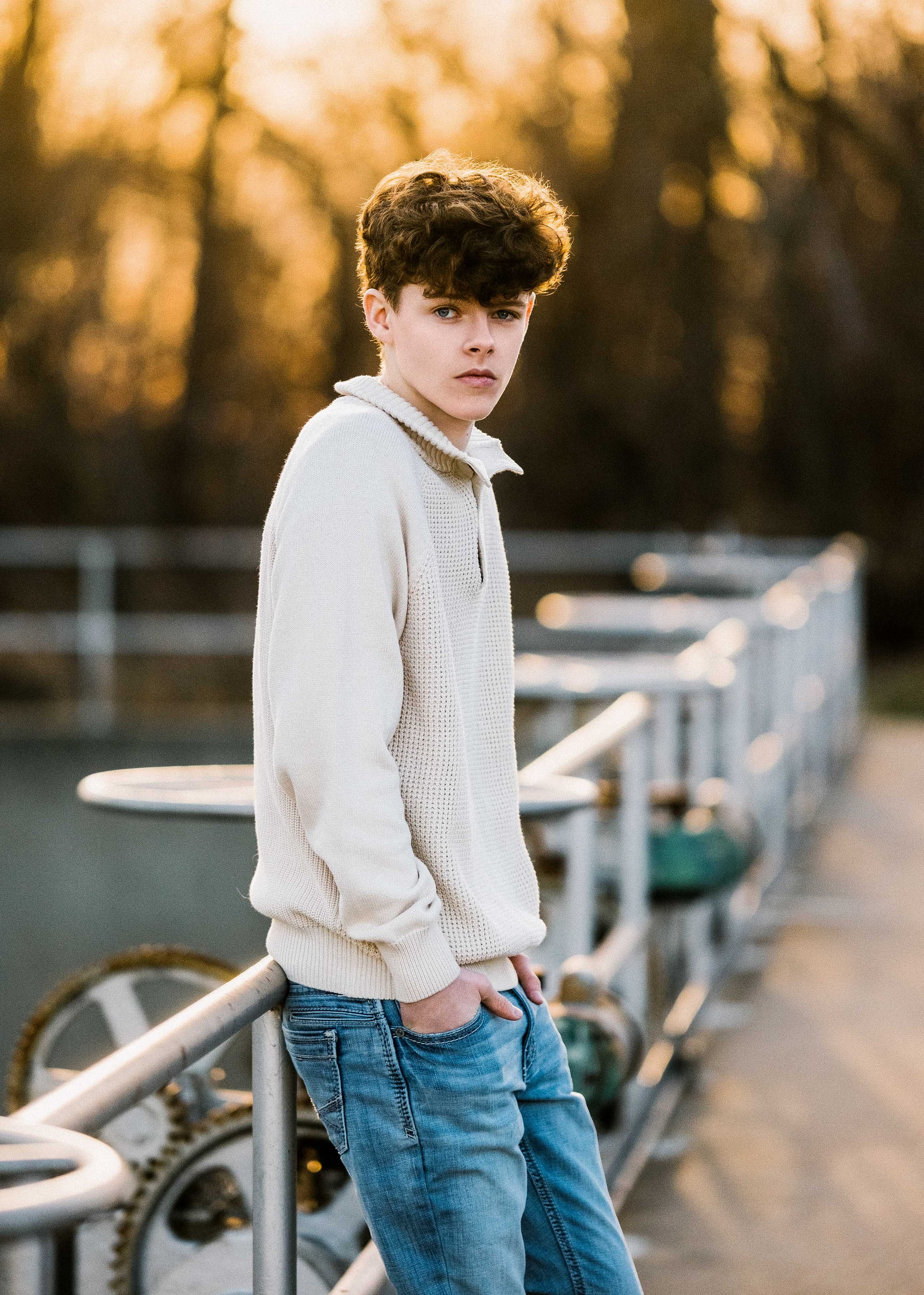 A young man with curly hair and fair skin leaning against a metal railing outdoors in warm, golden light. He is wearing a cream-colored knit sweater and blue jeans, with hands in his pockets, and looking at the camera. The background features blurred