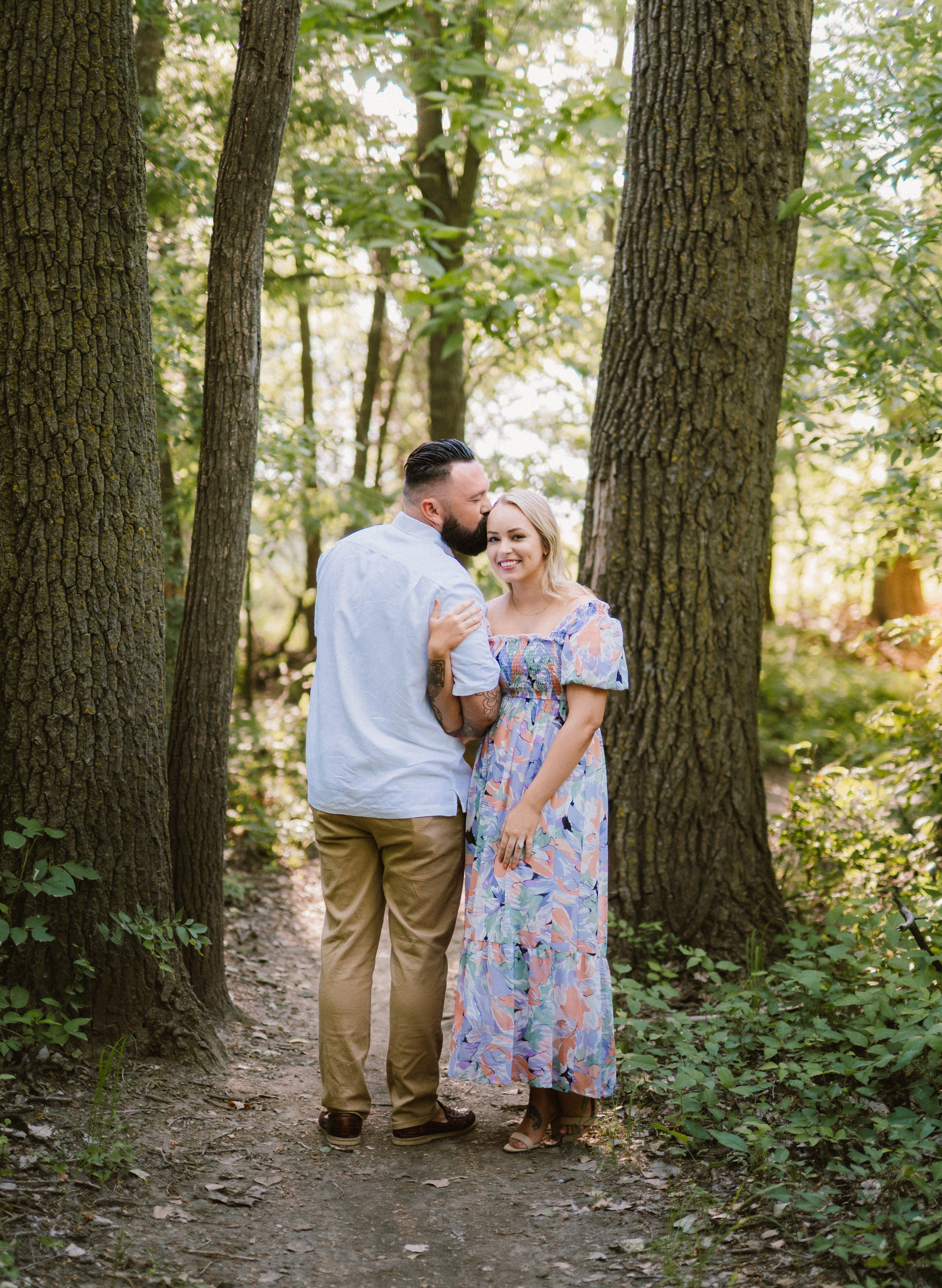 A couple standing on a forest trail, surrounded by tall trees and greenery, sharing a joyful moment with the man kissing the woman's forehead and both smiling.