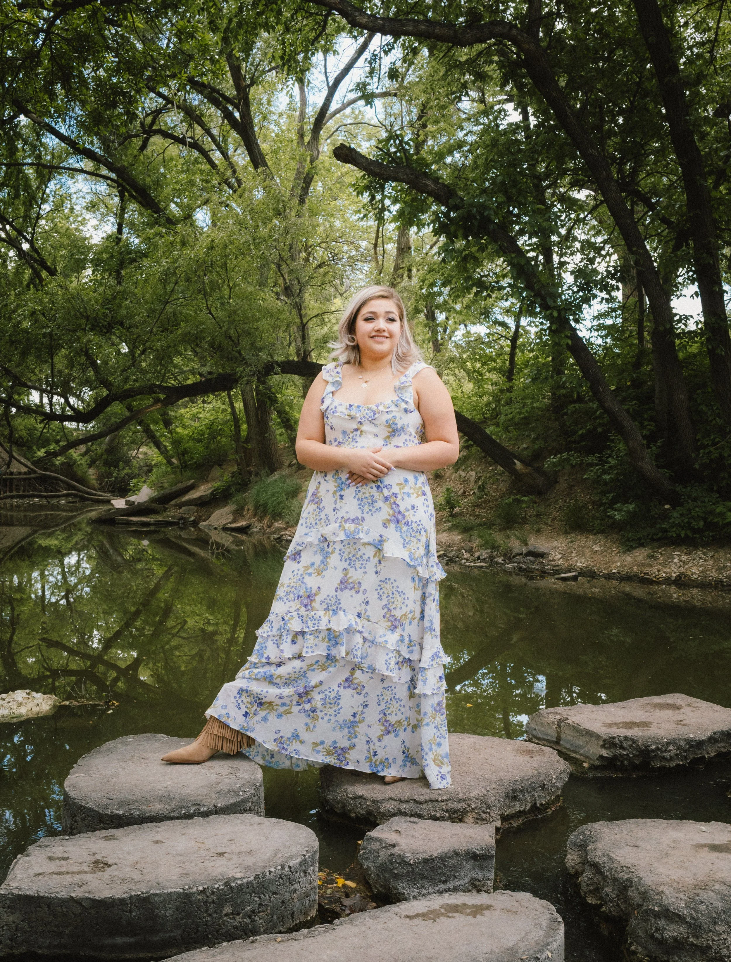A woman standing on large rocks in a shallow stream in a wooded area with dense green trees, wearing a long floral dress and boots.