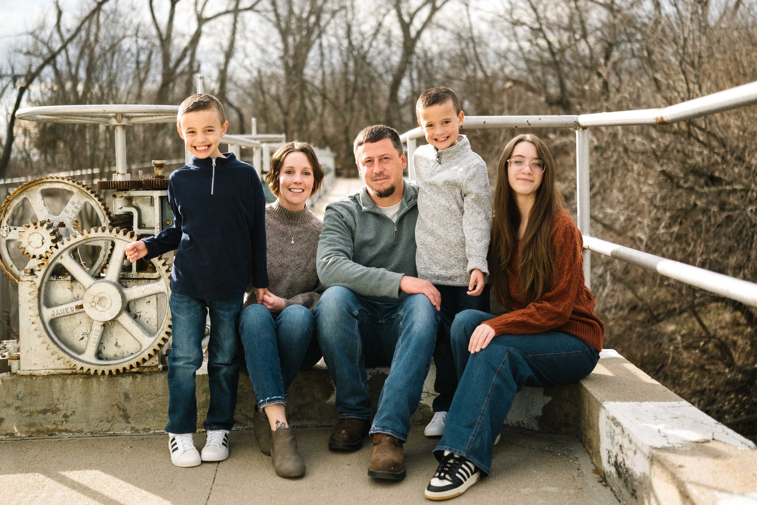 Family of six posing outdoors on a bridge with leafless trees in the background. Two boys, a woman, a man, a girl, and a young boy together smiling at the camera.