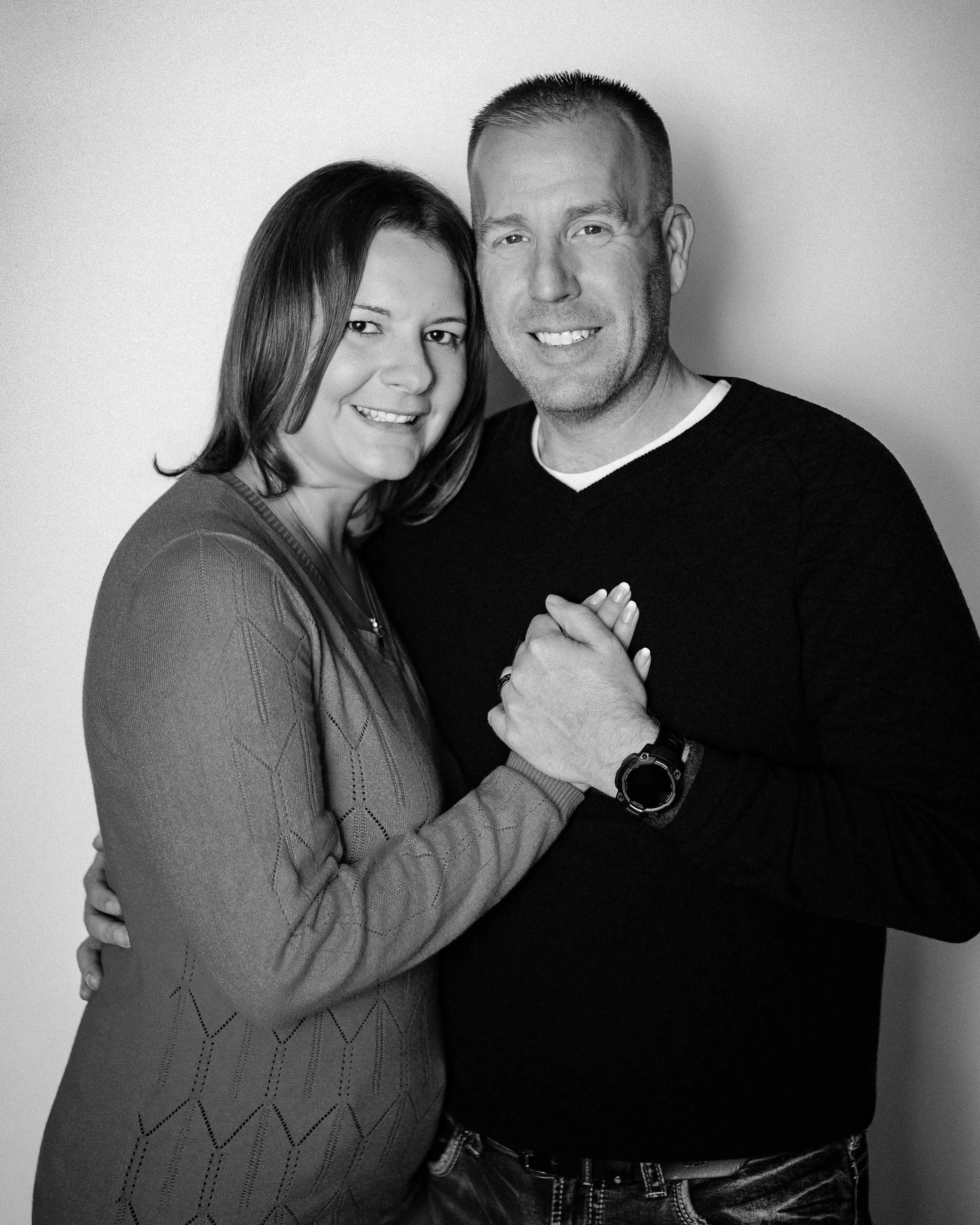 A black and white photo of a smiling couple holding hands, standing close together, facing the camera against a plain backdrop.