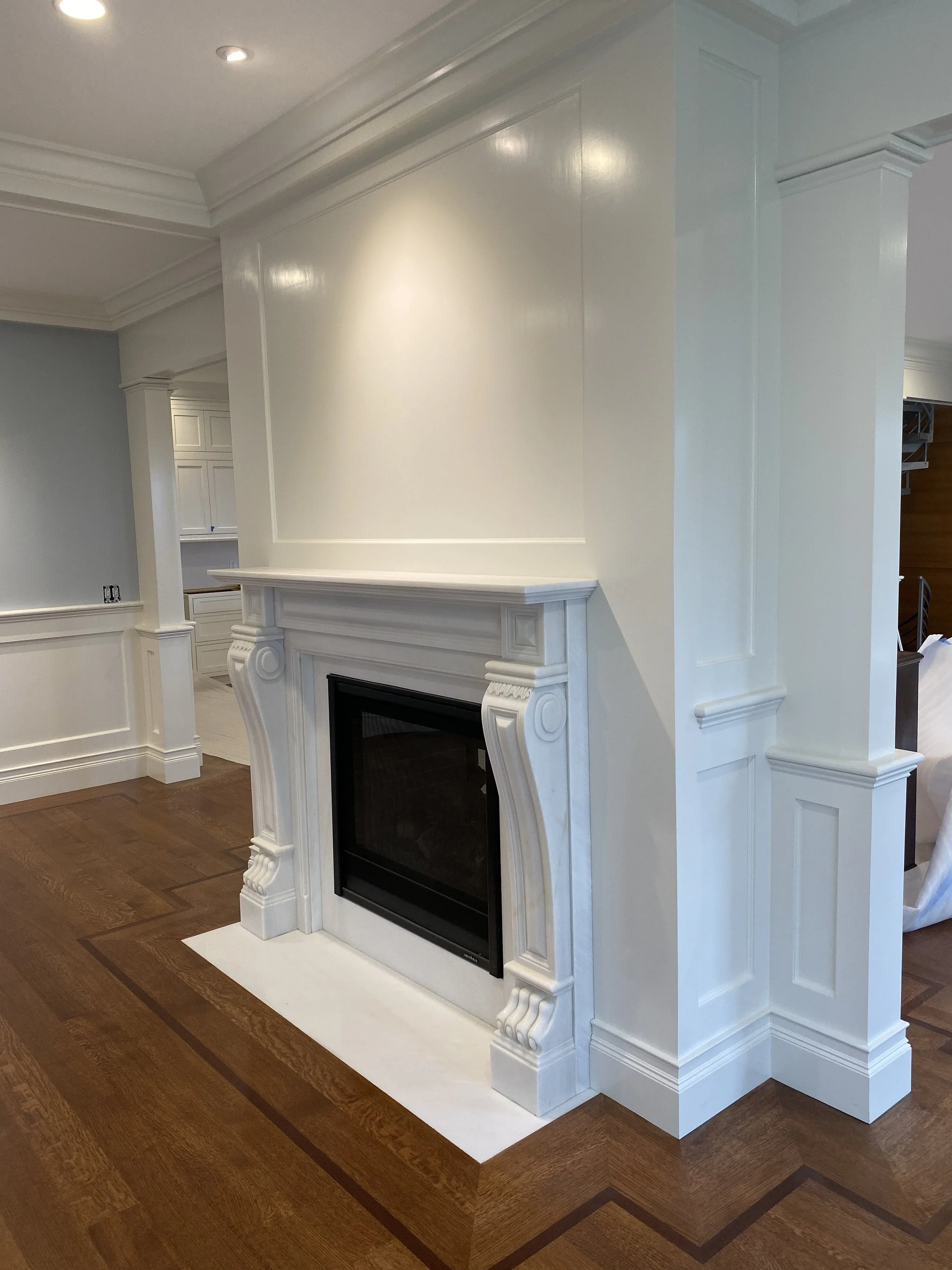 Interior view of a living room with a white fireplace surrounded by ornate molding and trim, hardwood floors, recessed lighting, and a corner with partial view of a staircase.