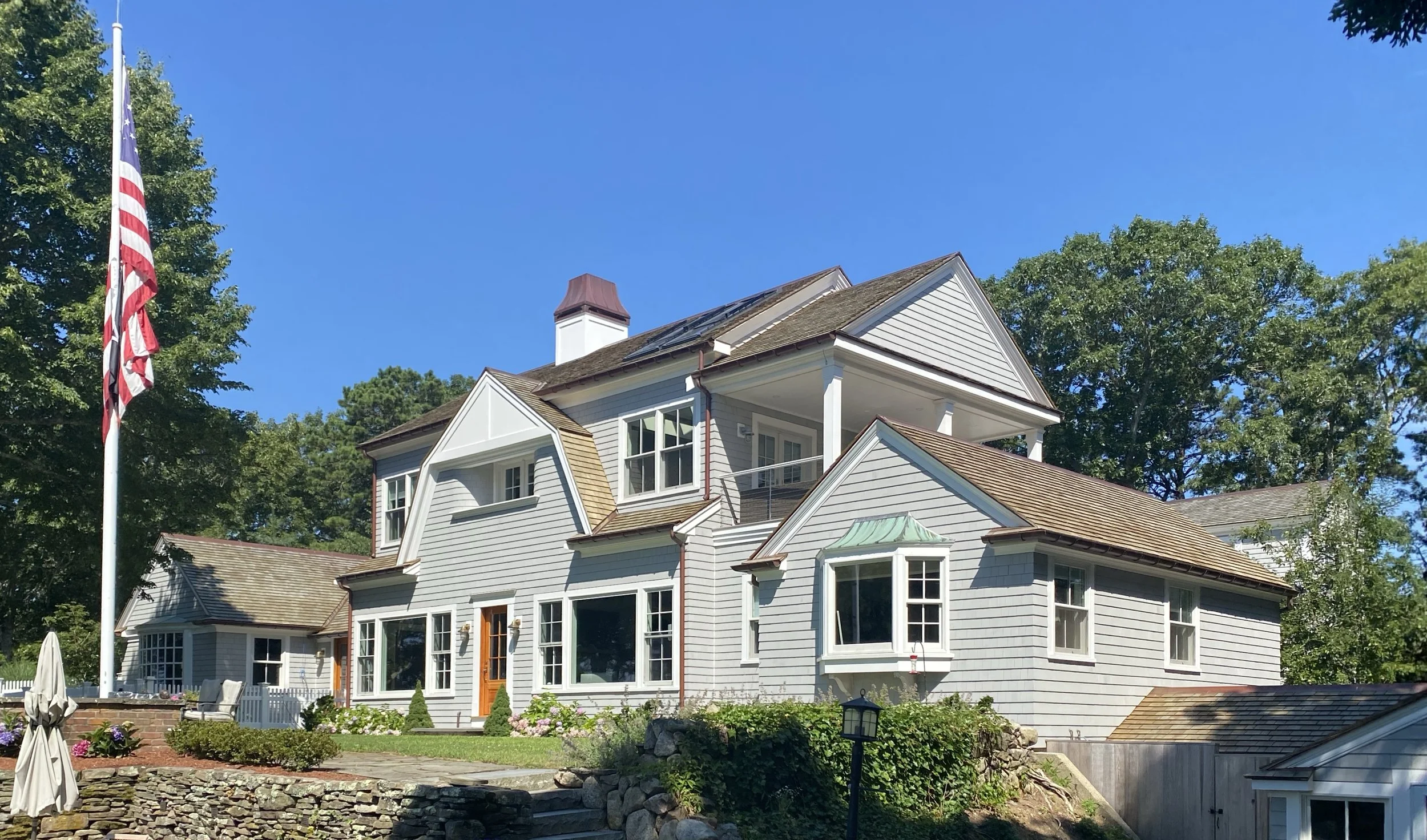 Large traditional house with white siding, several windows, a shingled roof, and a front porch. There is an American flag on a flagpole to the left, surrounded by greenery and landscaping.