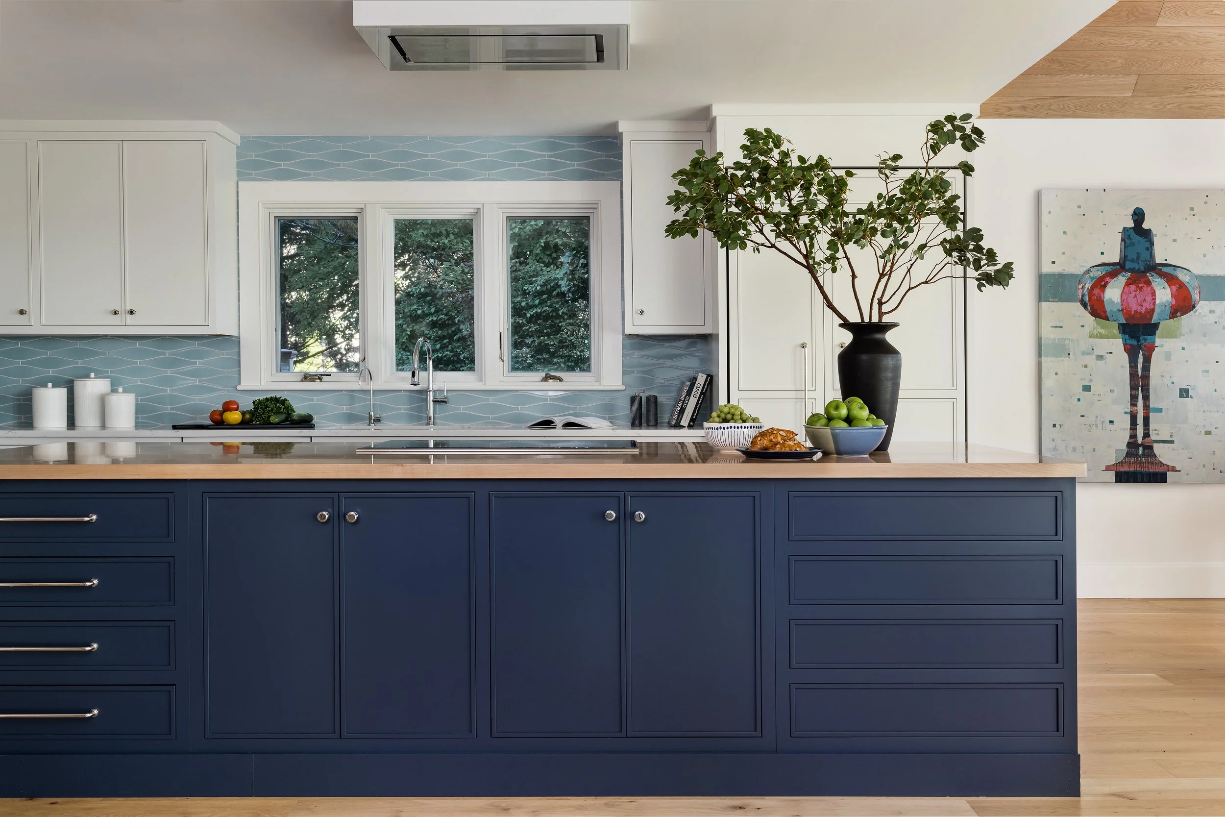 Modern kitchen with blue island, white cabinets, blue backsplash, and large window with view of trees.