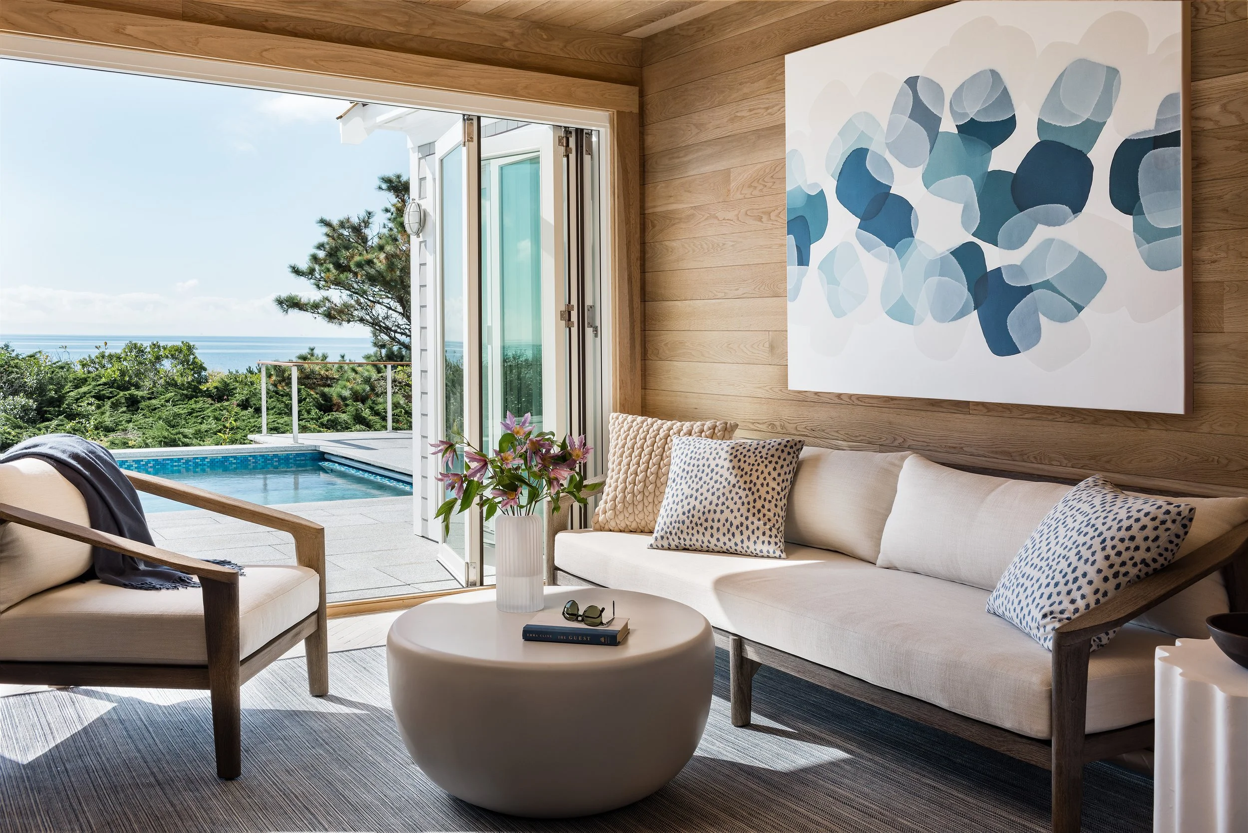 Living room with large glass door opening to outdoor pool and scenic ocean view, contemporary furniture, and abstract blue wall art.