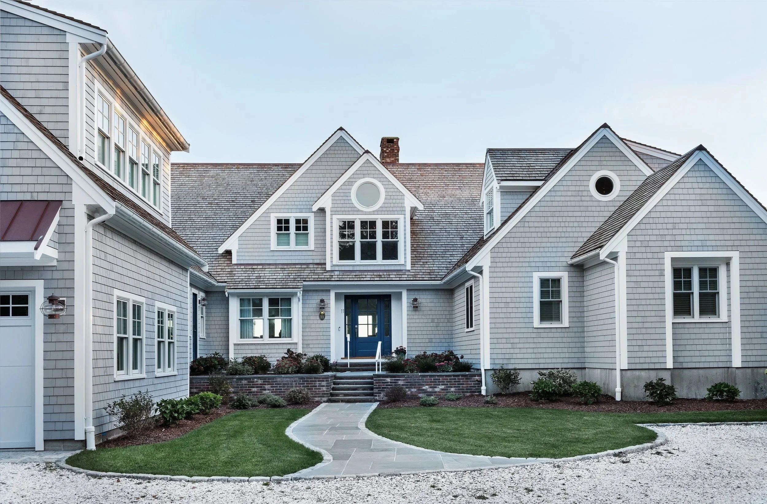 Front view of a large, classic American home with grey shingle siding, multiple gabled roofs, and white trim. Features include a prominent front porch with blue front door, small landscaped garden, and a curved stone pathway leading to the entrance.