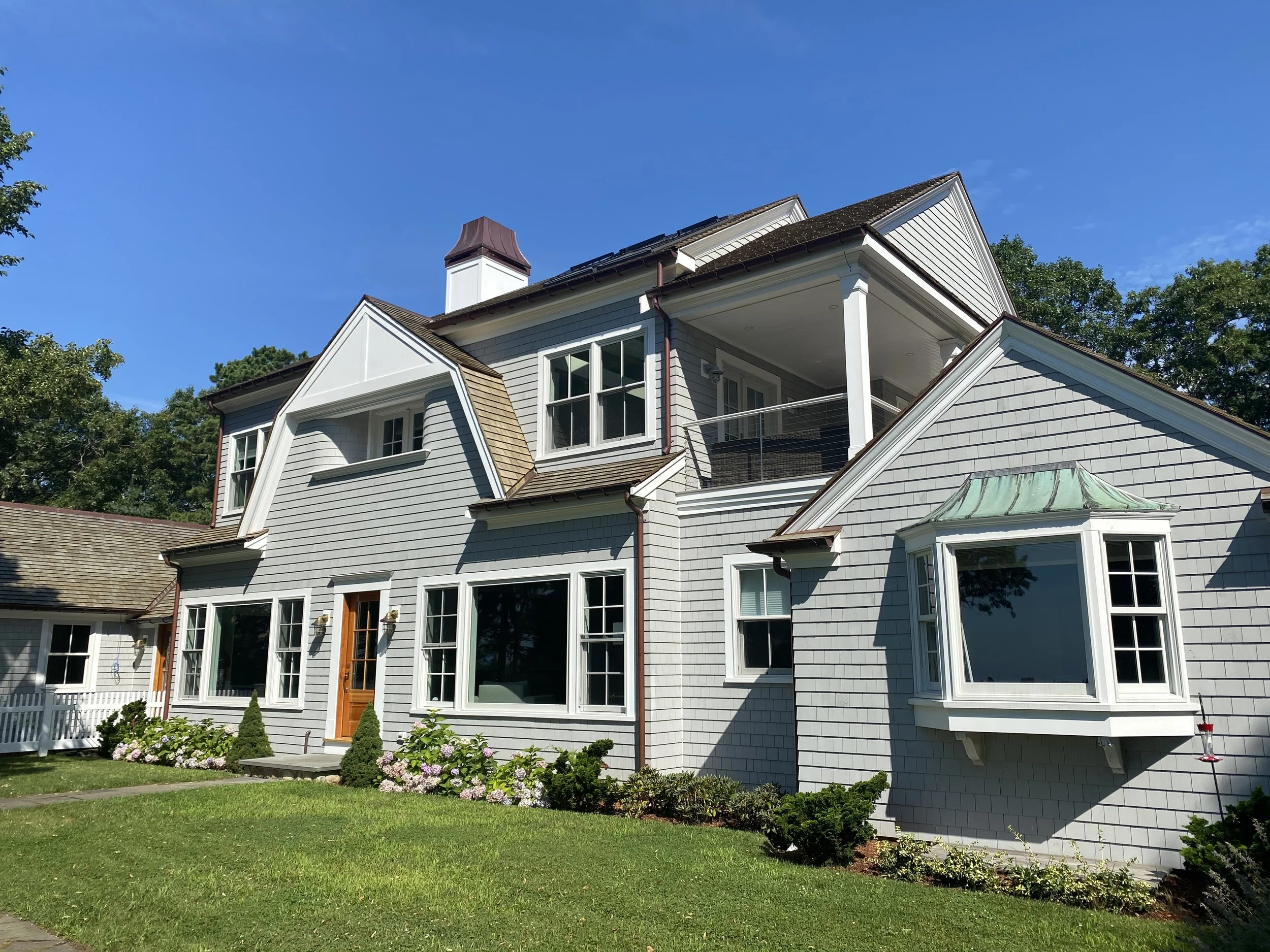 A large, multi-story house with white siding, multiple windows, a small balcony, and a landscaped front yard with green grass and bushes under a clear blue sky.