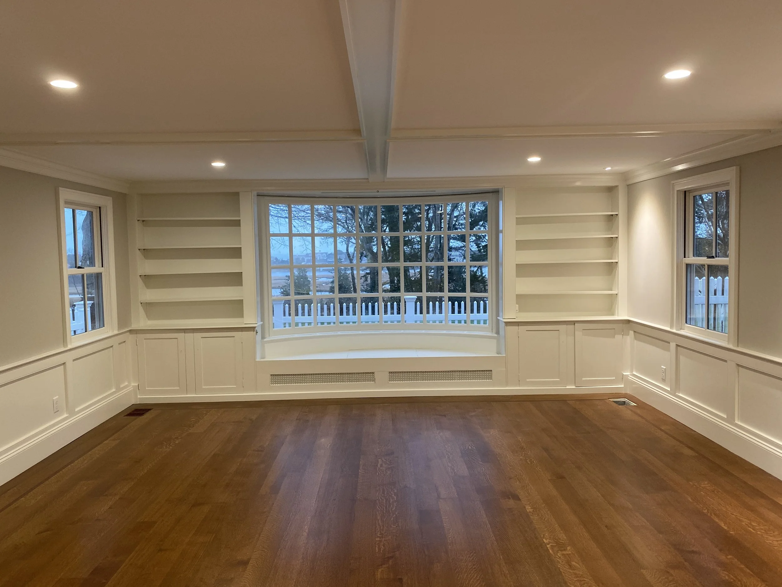 Empty living room with large bay window, built-in bookshelves on both sides, hardwood flooring, and recessed ceiling lights.