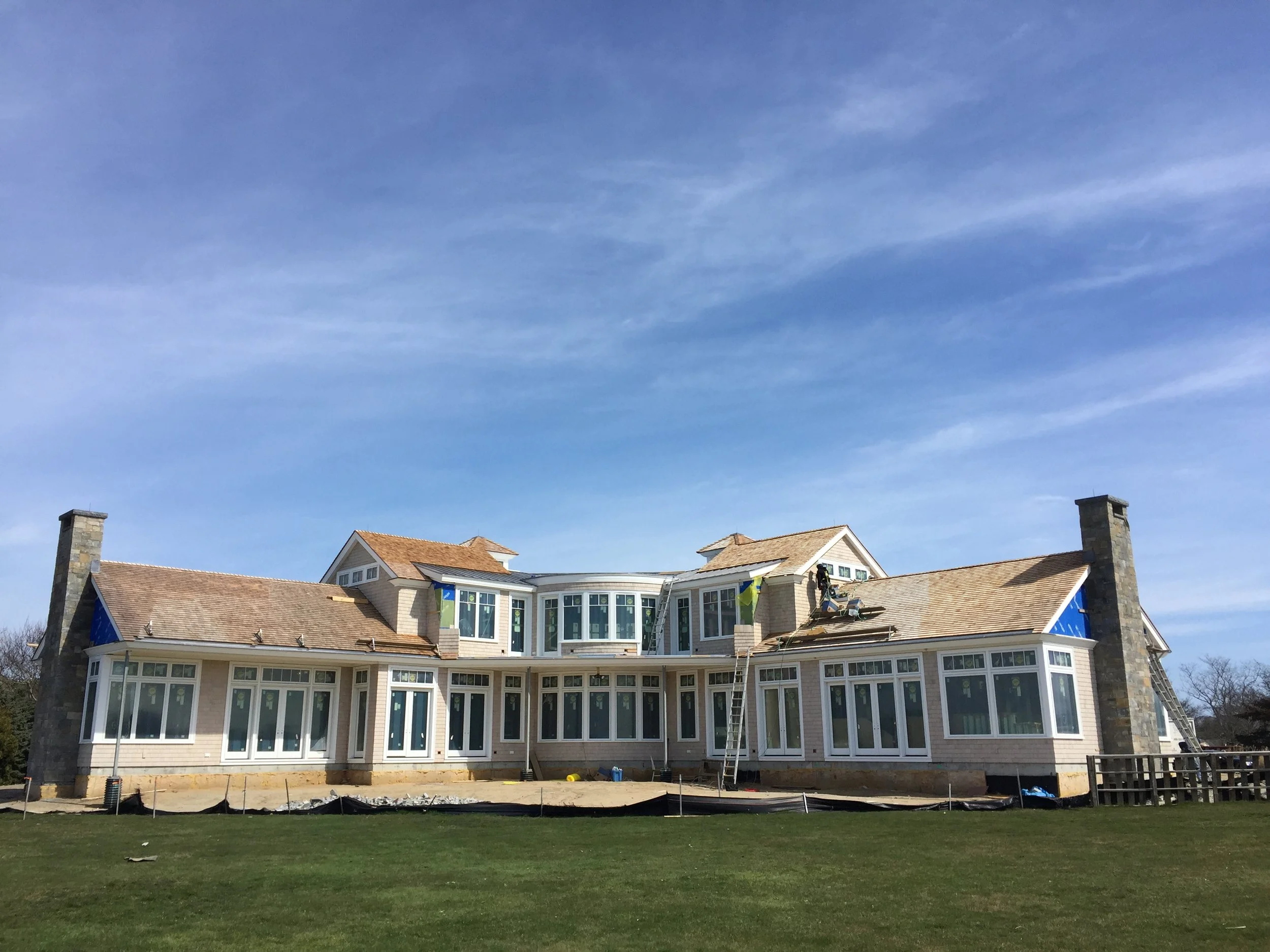 Large house under construction with scaffolding and workers on the roof, surrounded by a grassy lawn and a blue sky.