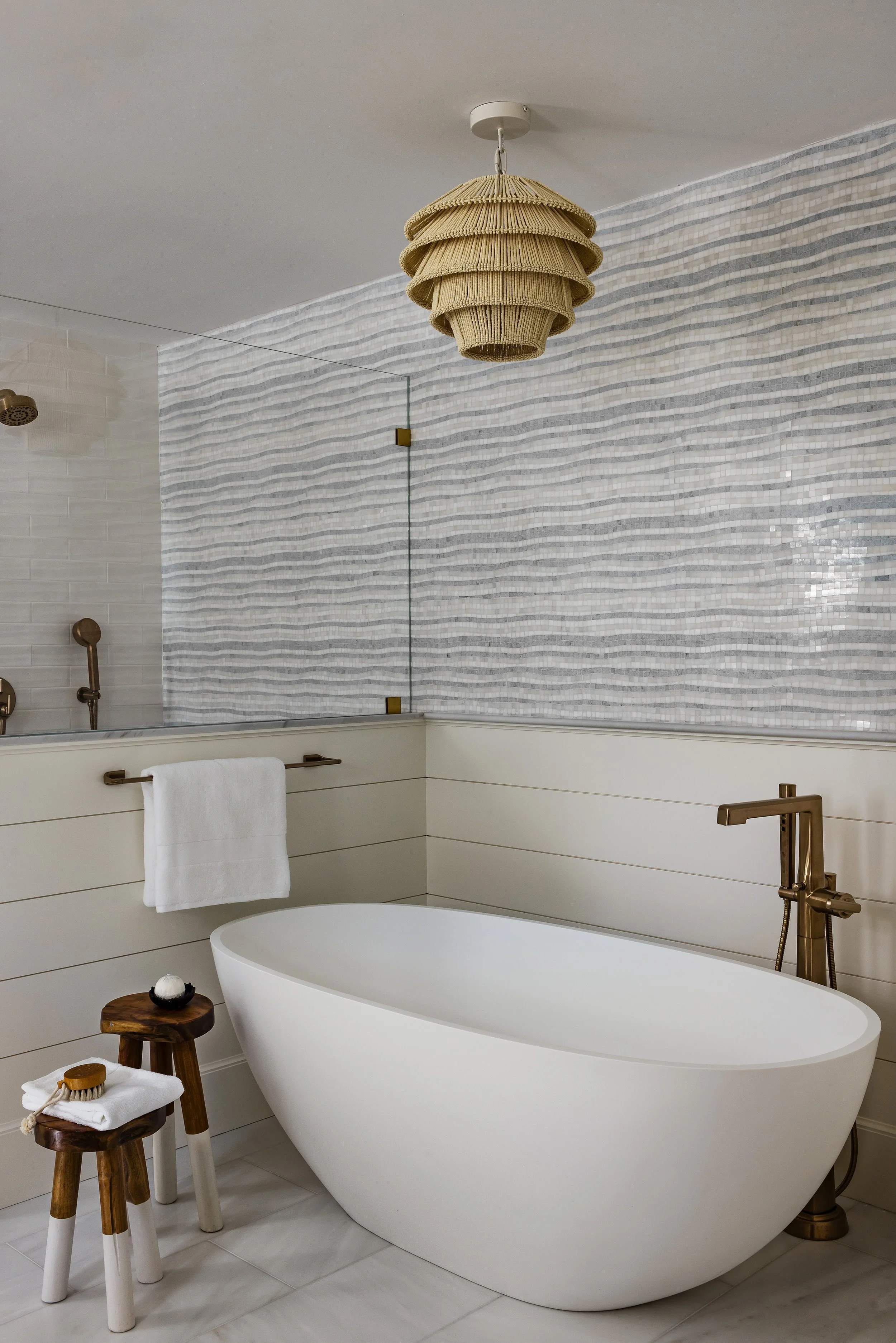 Modern bathroom with a white freestanding bathtub, gold fixtures, a towel hanging on a brass bar, two small wooden stools, a woven pendant light, and a textured mosaic tile wall.