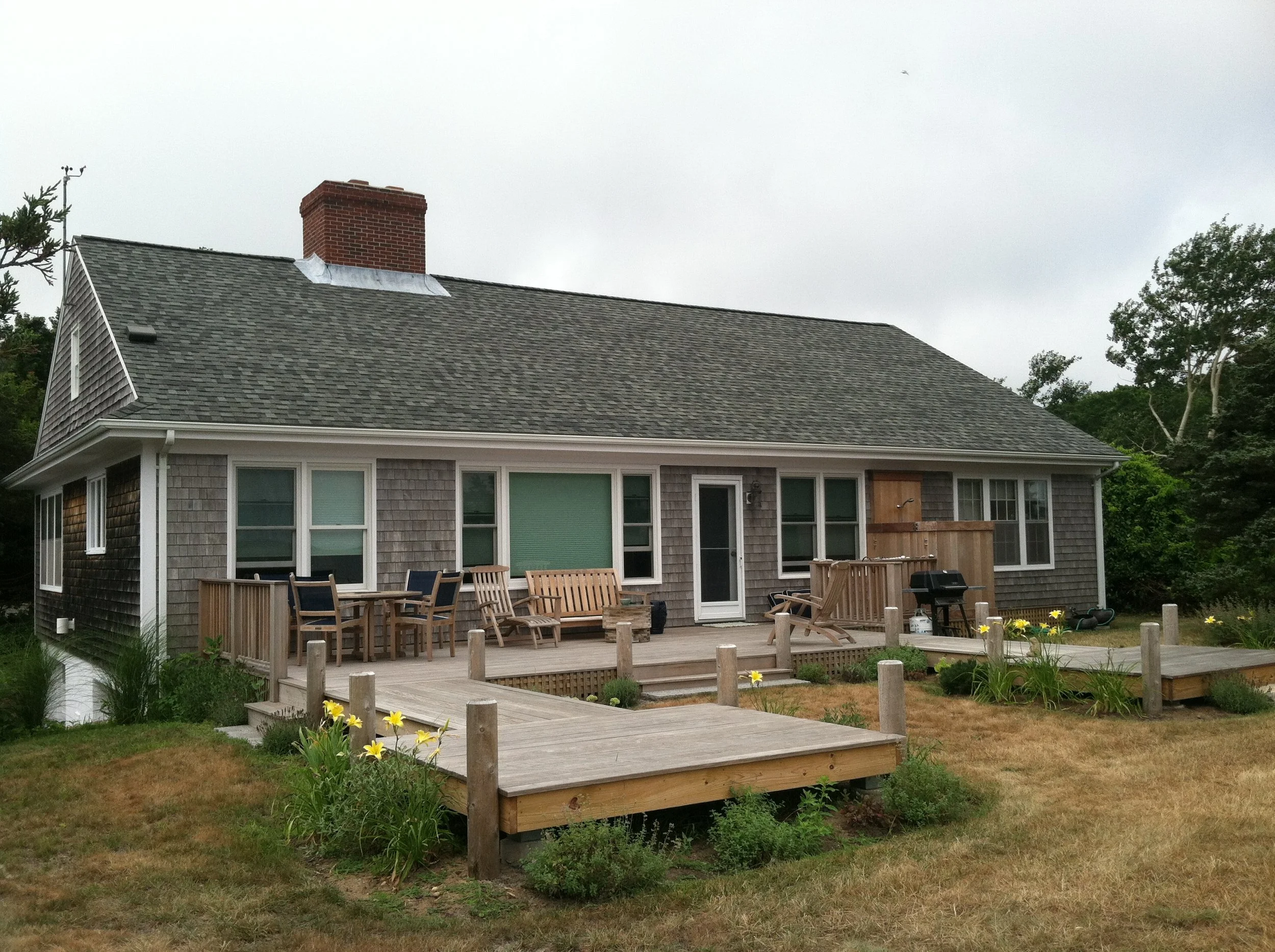 Backyard view of a house with a wooden deck, outdoor furniture, and a grill, surrounded by garden plants and trees under an overcast sky.