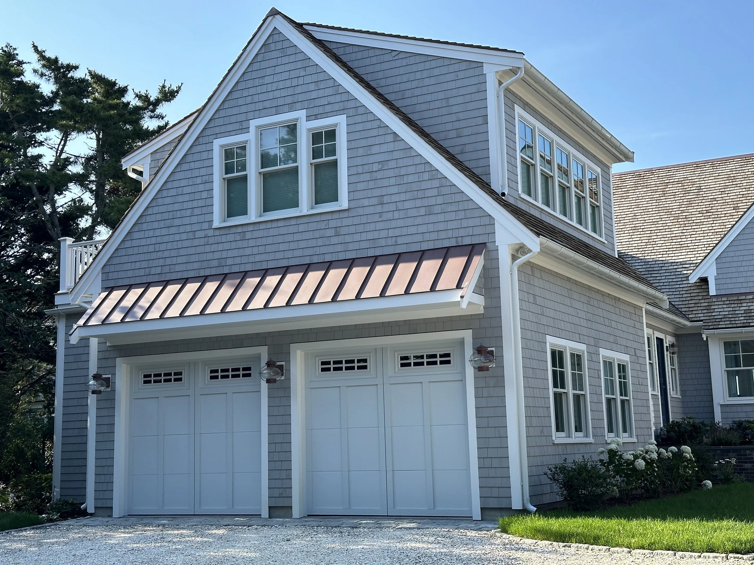 A two-story house with gray shingle siding, white window trims, a brown metal awning over the garage, and a gravel driveway.
