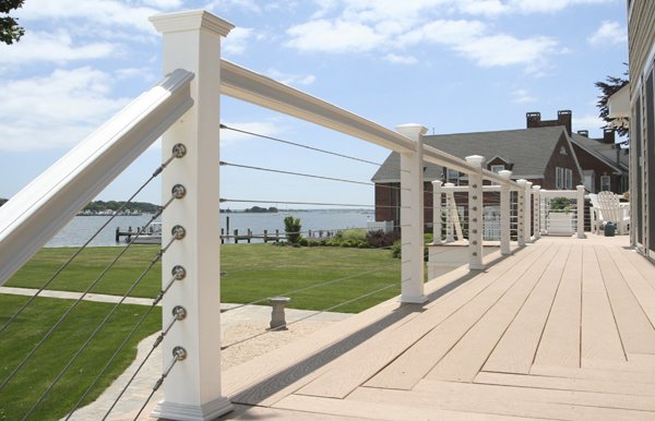 A spacious wooden deck with white railing overlooking a lake, with a house nearby, under a partly cloudy sky.