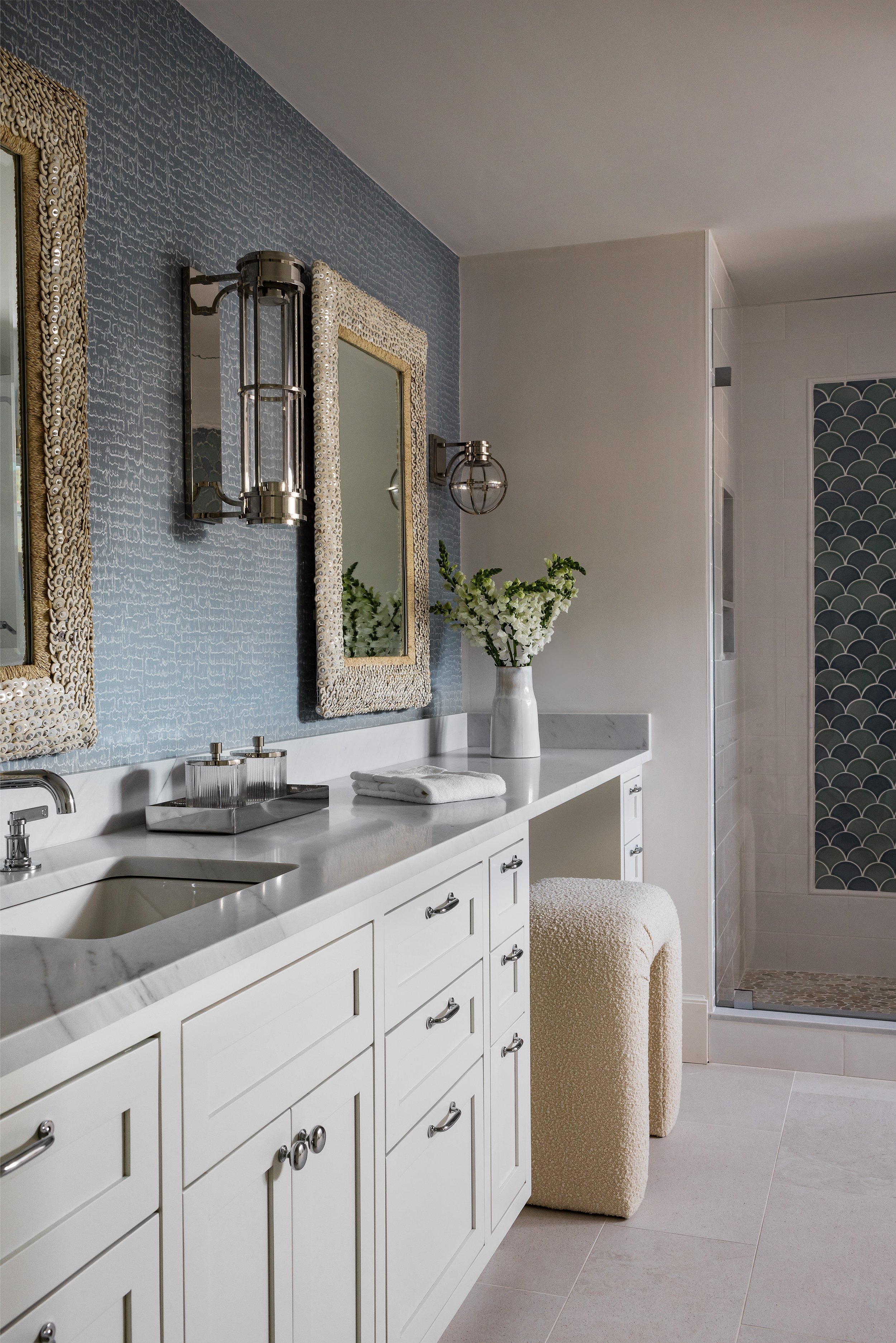 A modern bathroom with white cabinetry, a marble countertop, and a beige textured stool. There are two rectangular mirrors with ornate beige frames, a pair of sconces, and a white vase with white flowers. In the background, there's a walk-in shower w
