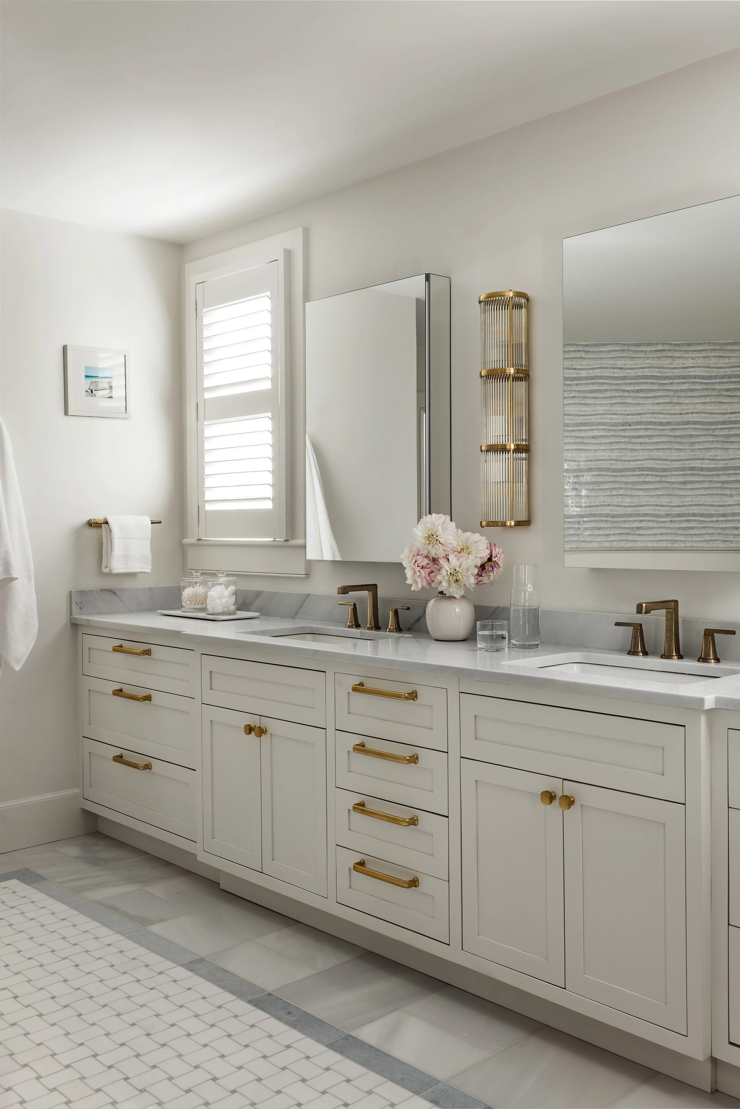 A modern white bathroom vanity with gold hardware, two sinks, and a marble countertop. There is a framed mirror, a floral vase, and a glass of water on the counter. Light enters through a small window with white shutters, and a towel hangs on a gold 