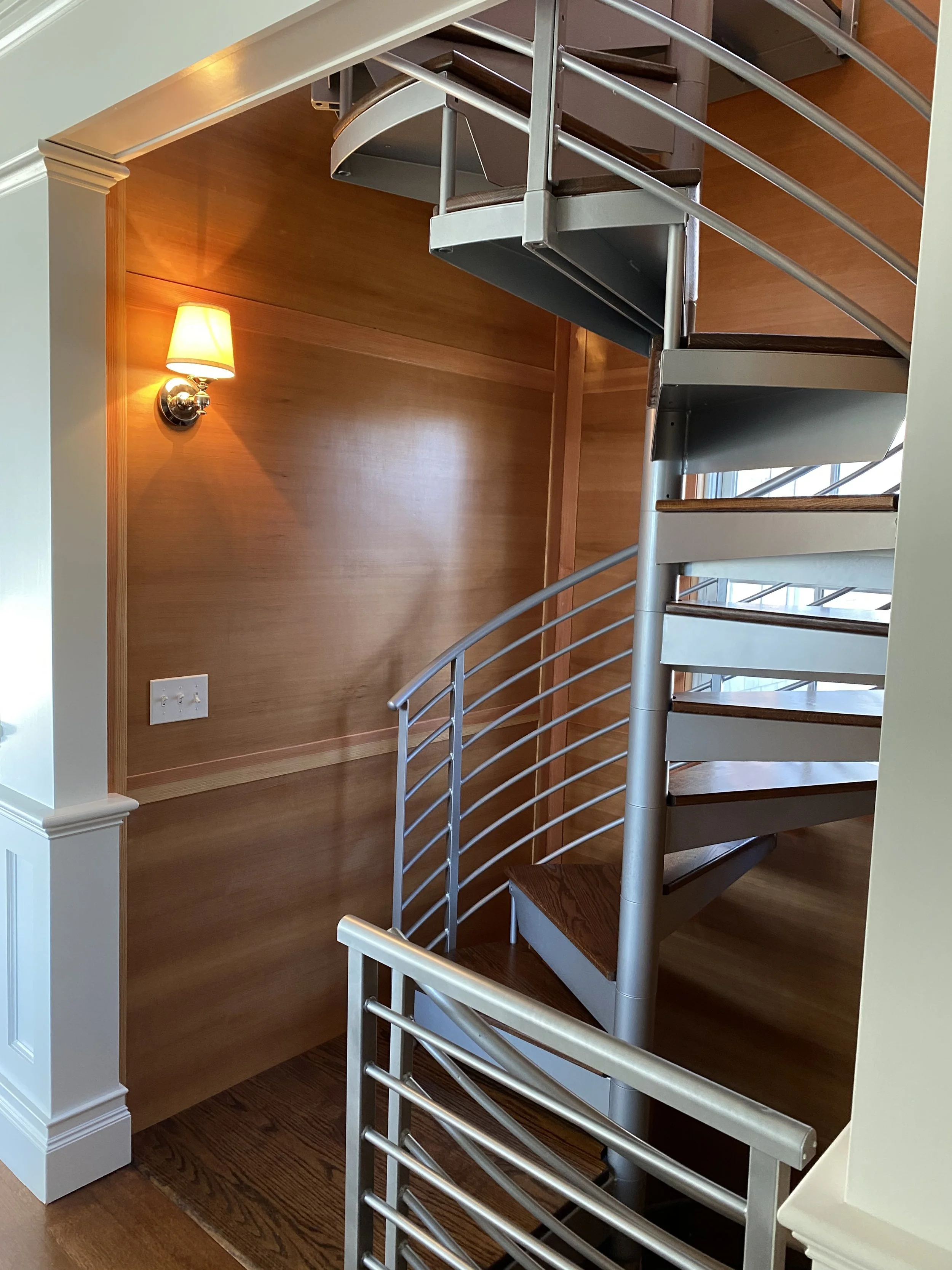 Interior view of a spiral staircase with metal railings next to a wood-paneled wall and a wall-mounted lamp