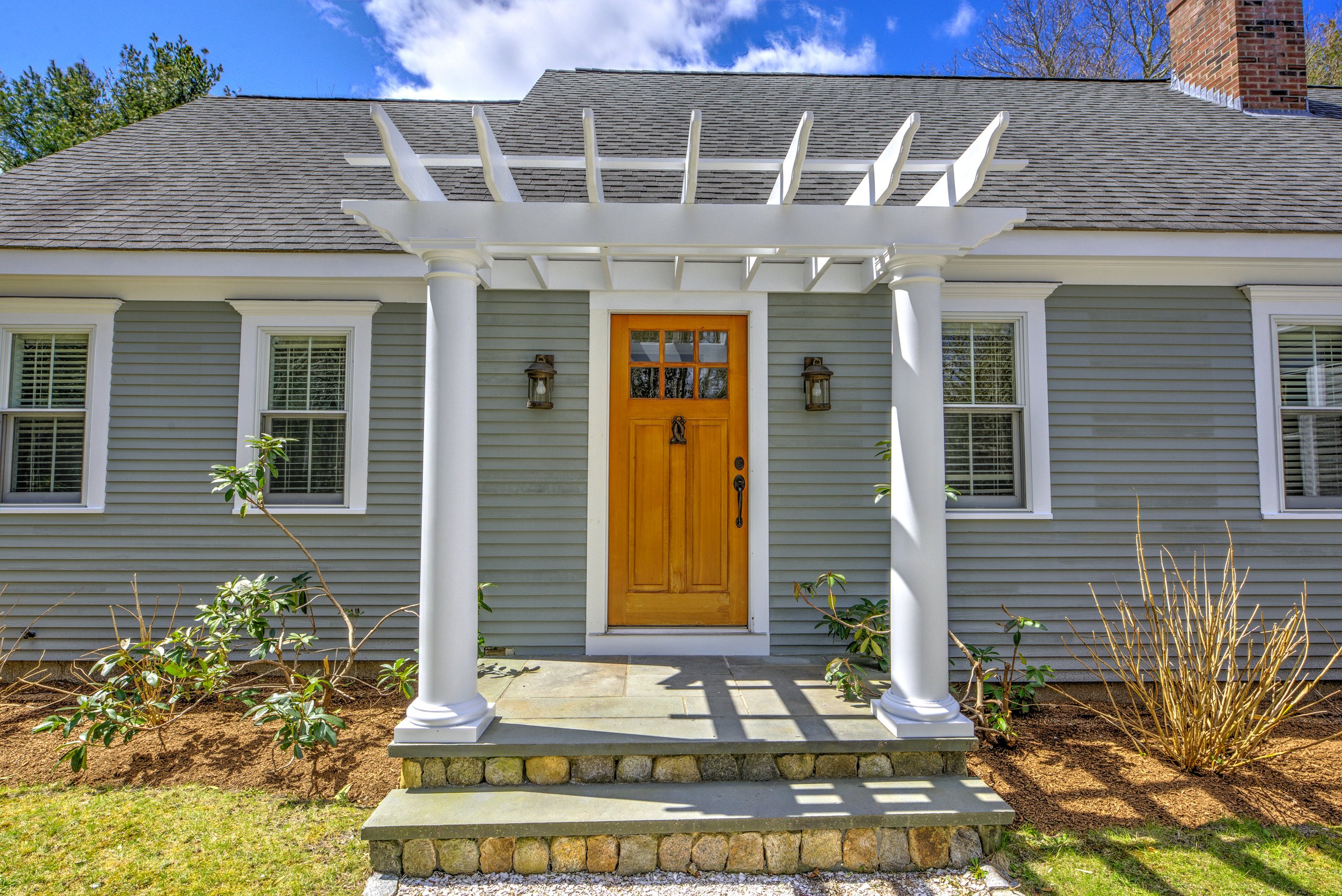 Front view of a house entrance with a wooden door framed by white columns supporting a white pergola. There are two windows on either side of the door, garden plants, a small set of stairs, and a brick chimney on the roof under a partly cloudy sky.