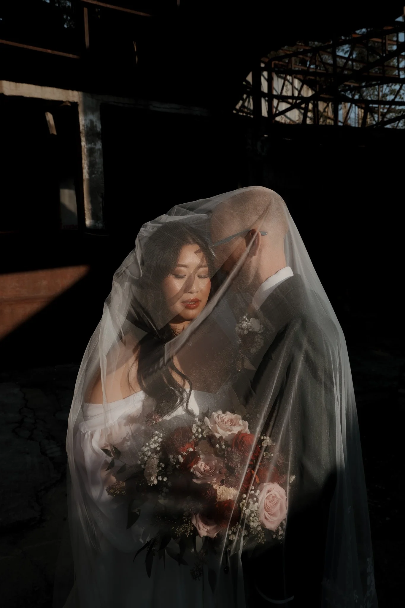 A bride and groom share a intimate moment under a sheer veil, with the bride holding a bouquet of pink and white roses, in a dark outdoor setting.