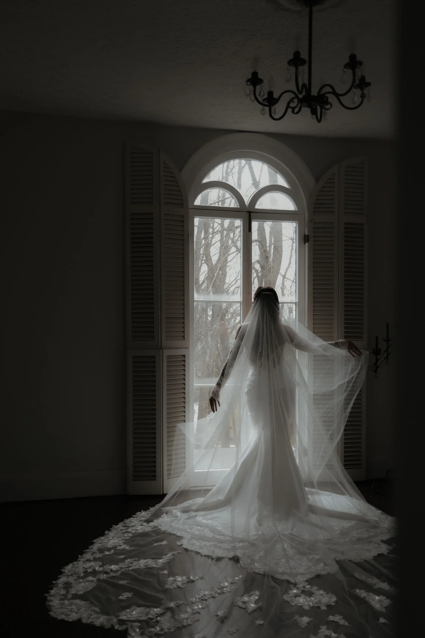 A bride in a wedding dress with a long train and veil standing in front of a large window with shutters, letting in soft natural light, in an elegant room with a chandelier on the ceiling.