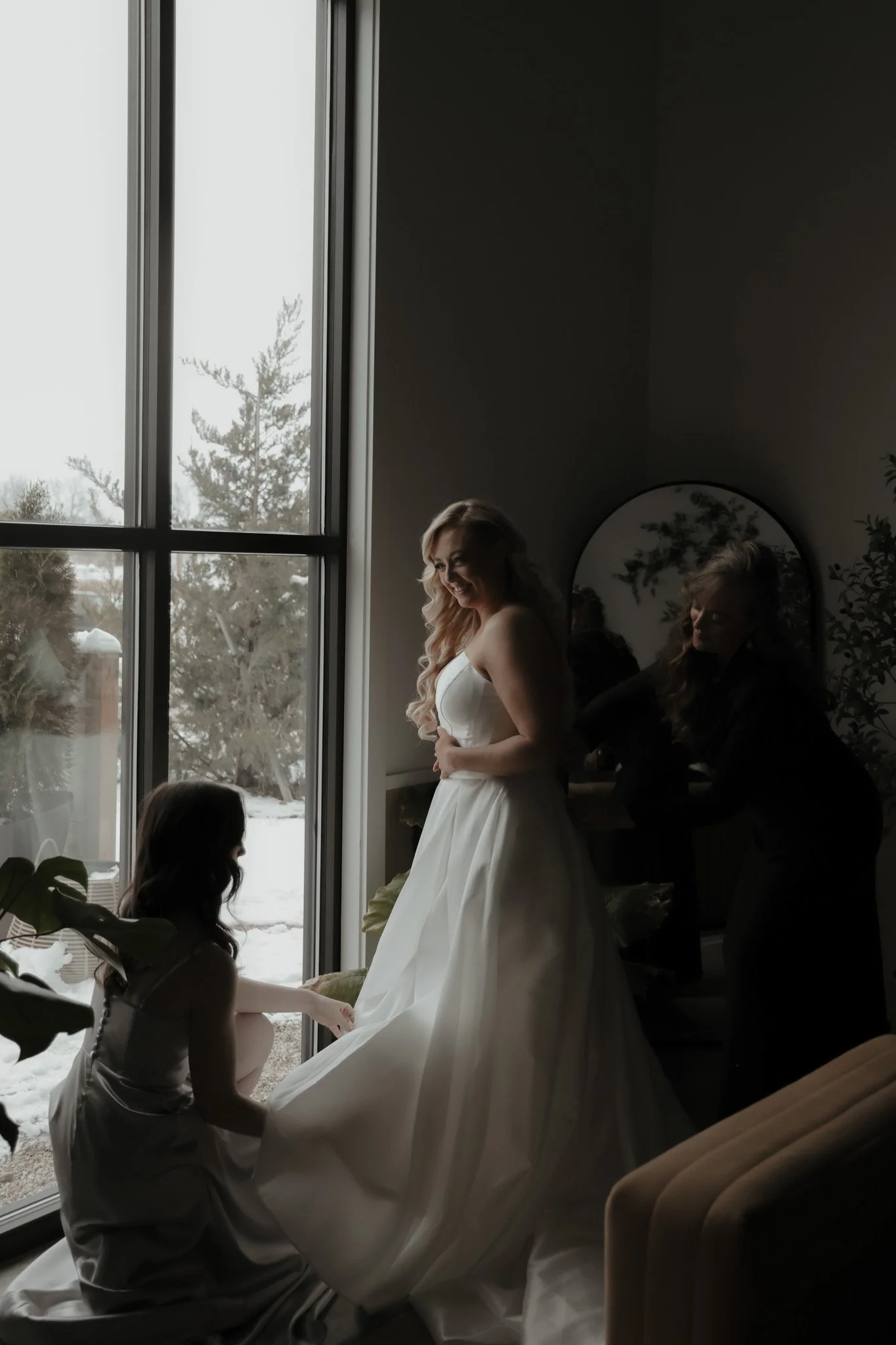 A bride in a white wedding gown is smiling while standing indoors near a large window. A girl in a dress is adjusting the gown's train, and a woman is helping her backstage. Outside the window, snow and trees are visible.