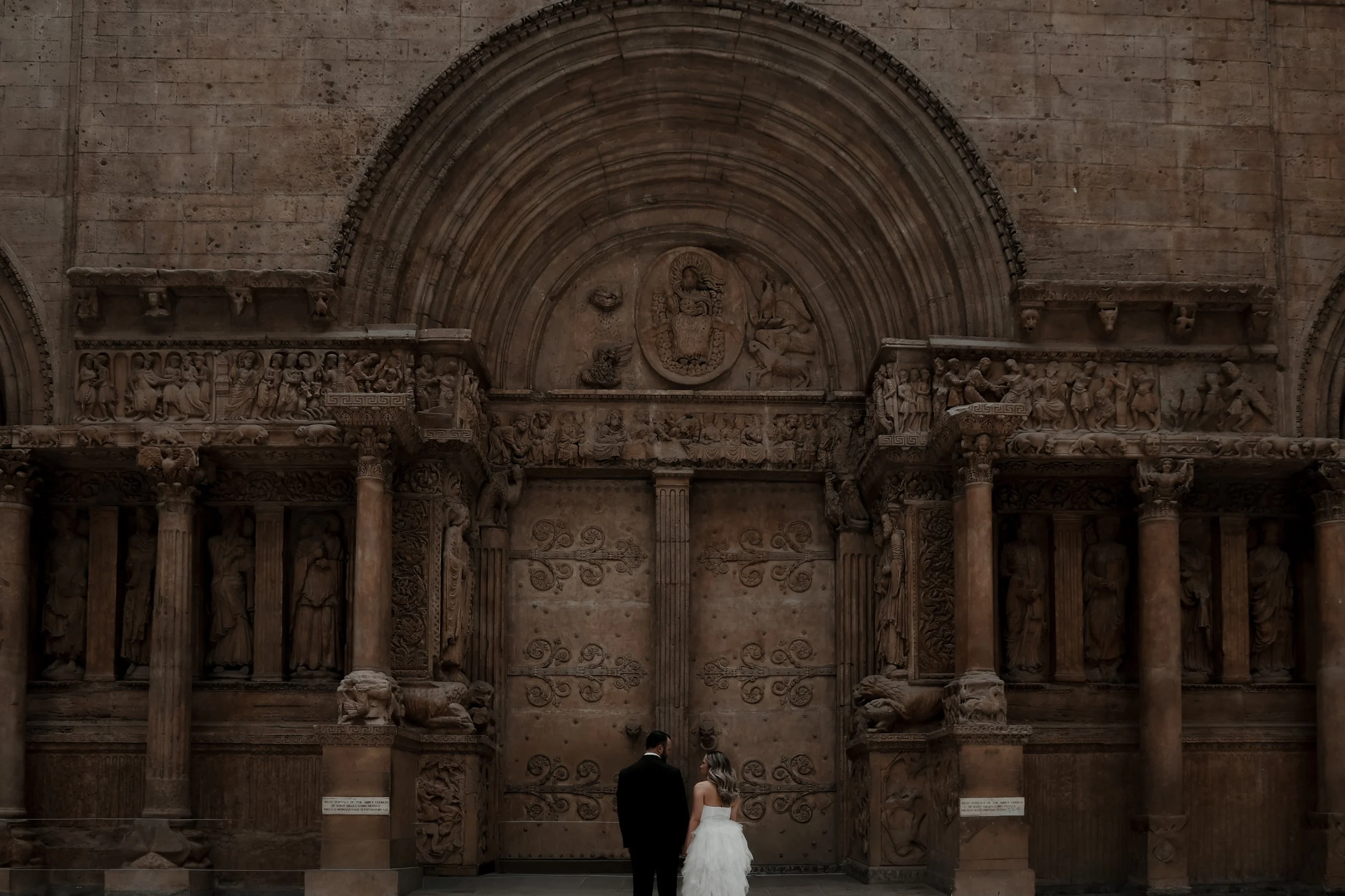 A bride and groom in wedding attire standing in front of an ornate ancient stone door with intricate carvings and columns.