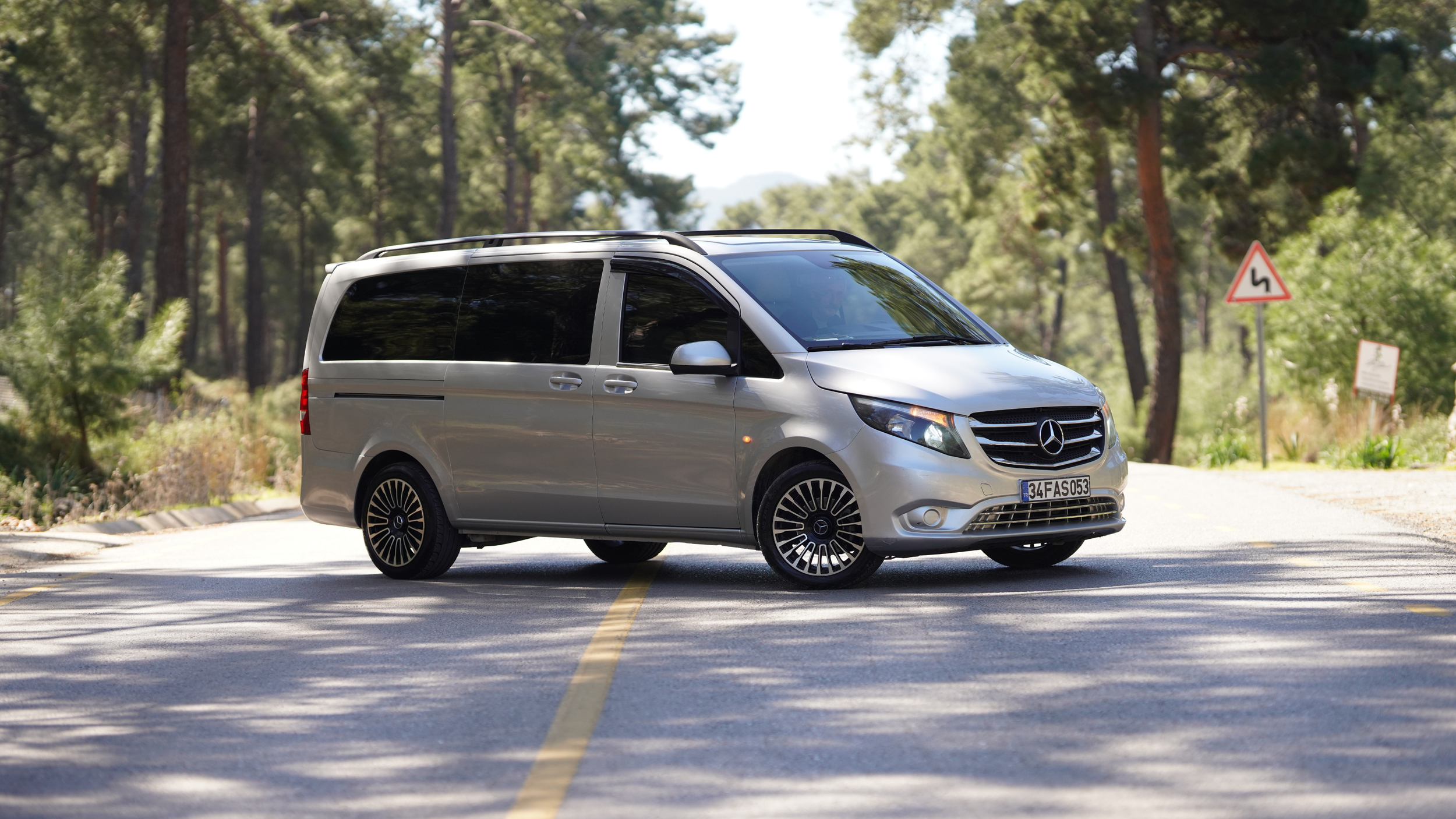 A silver Mercedes-Benz van stopped in the middle of a two-lane road surrounded by trees, with a road sign indicating winding road ahead in the background.