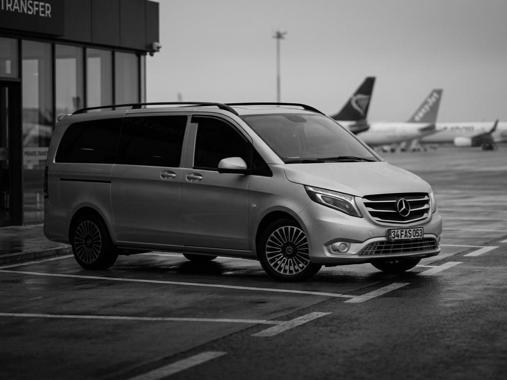 A silver Mercedes-Benz van parked at an airport near an airplane taxiway, with airplanes in the background.
