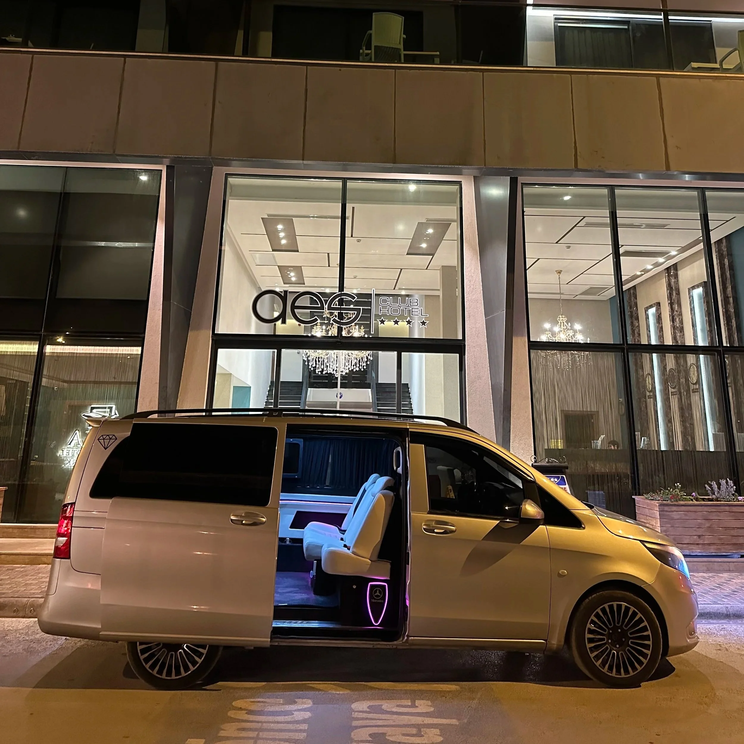 A beige Mercedes-Benz van parked outside a hotel at night, with one side door open revealing white interior seats and a purple illuminated logo, in front of a modern glass-fronted hotel entrance with a chandelier visible inside.