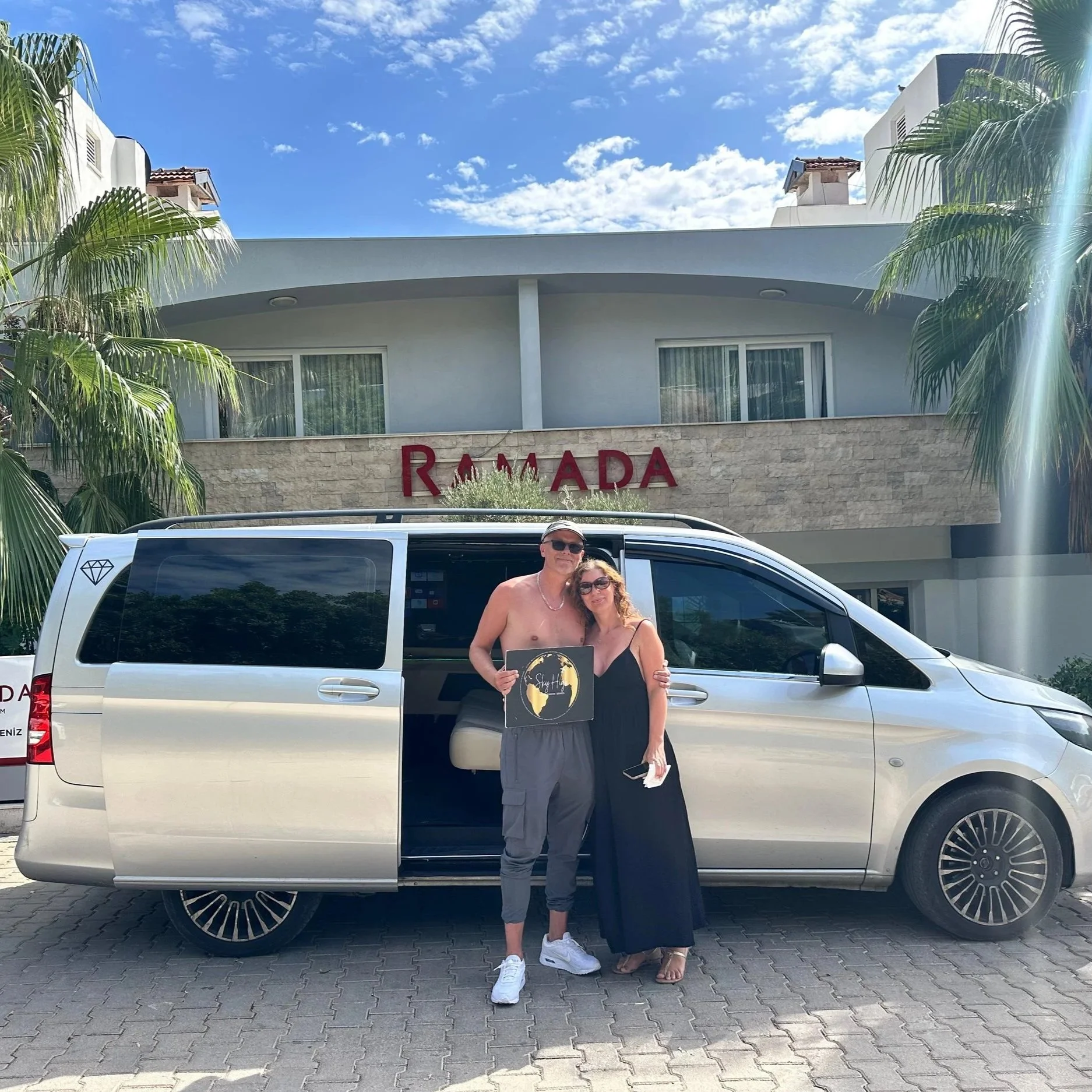 Couple standing beside a silver van in front of a hotel with a sign reading "Ramada", under a partly cloudy blue sky, with palm trees on either side.