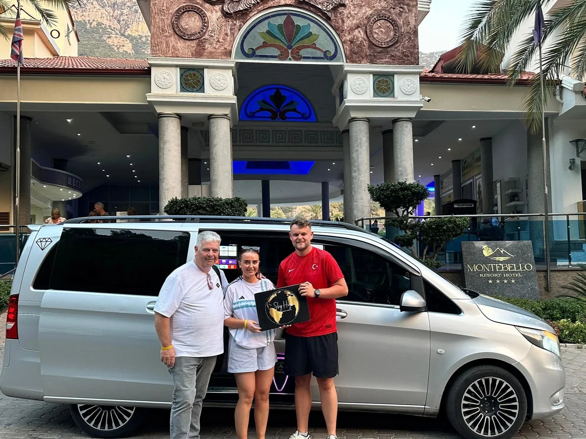 Three people stand in front of a silver van parked outside the Montebello Resort Hotel. The background features hotel entrance with tall columns, decorative architecture, and a sign for the hotel. One of the people is holding a black shopping bag.