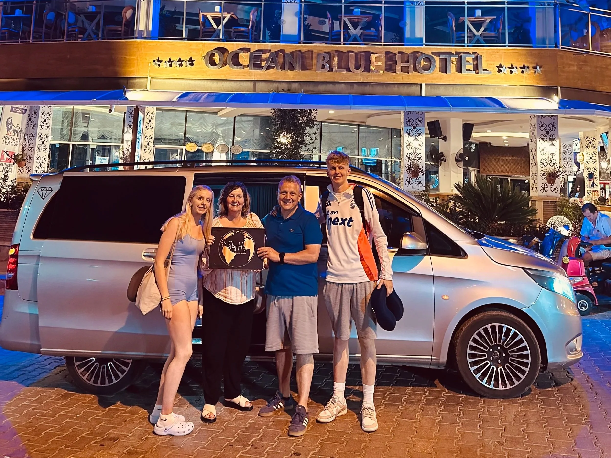 Four people standing in front of a silver van outside the Ocean Blue Hotel at night. The group is posing for the photo, with the hotel building illuminated in the background.