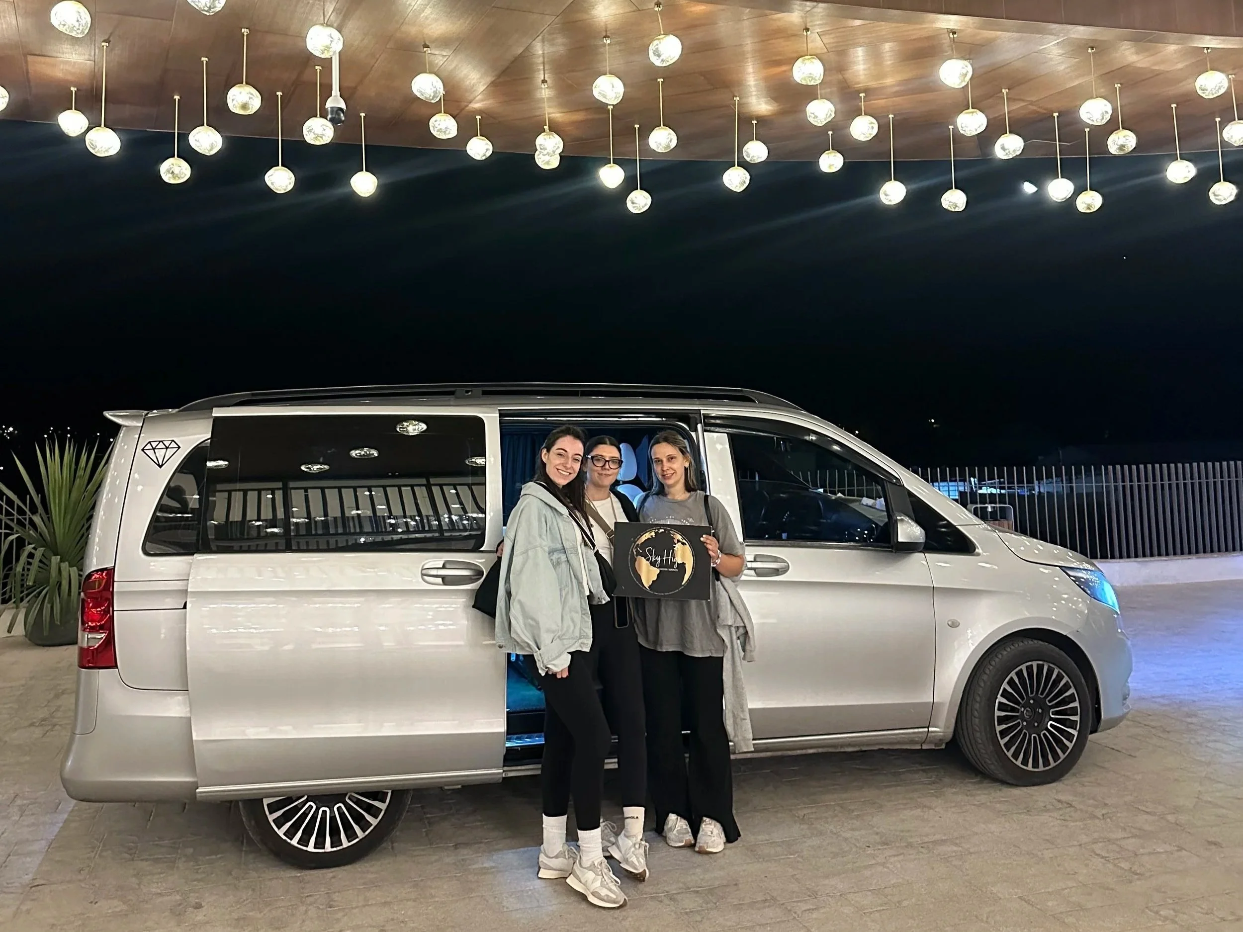 Three women stand next to a silver minivan at night, one of them holding a black sign with a gold logo. The scene is illuminated by hanging globe lights above.