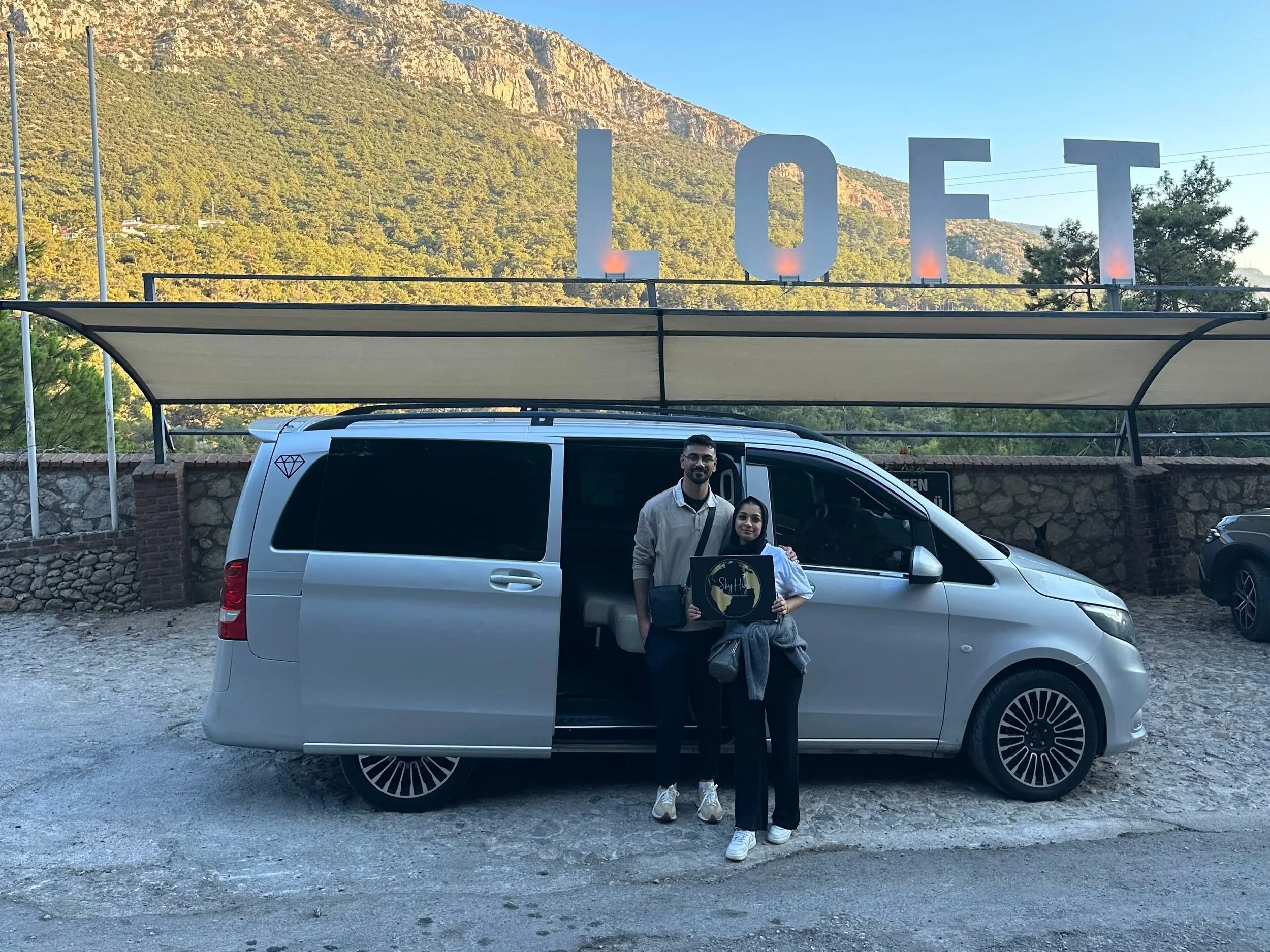 Two people standing in front of a silver van, posing for a photo. The person on the left is a man with a beard, wearing glasses and a light-colored jacket. The person on the right is a woman holding a sign that says 'Sky Hill.' Behind them, there is 