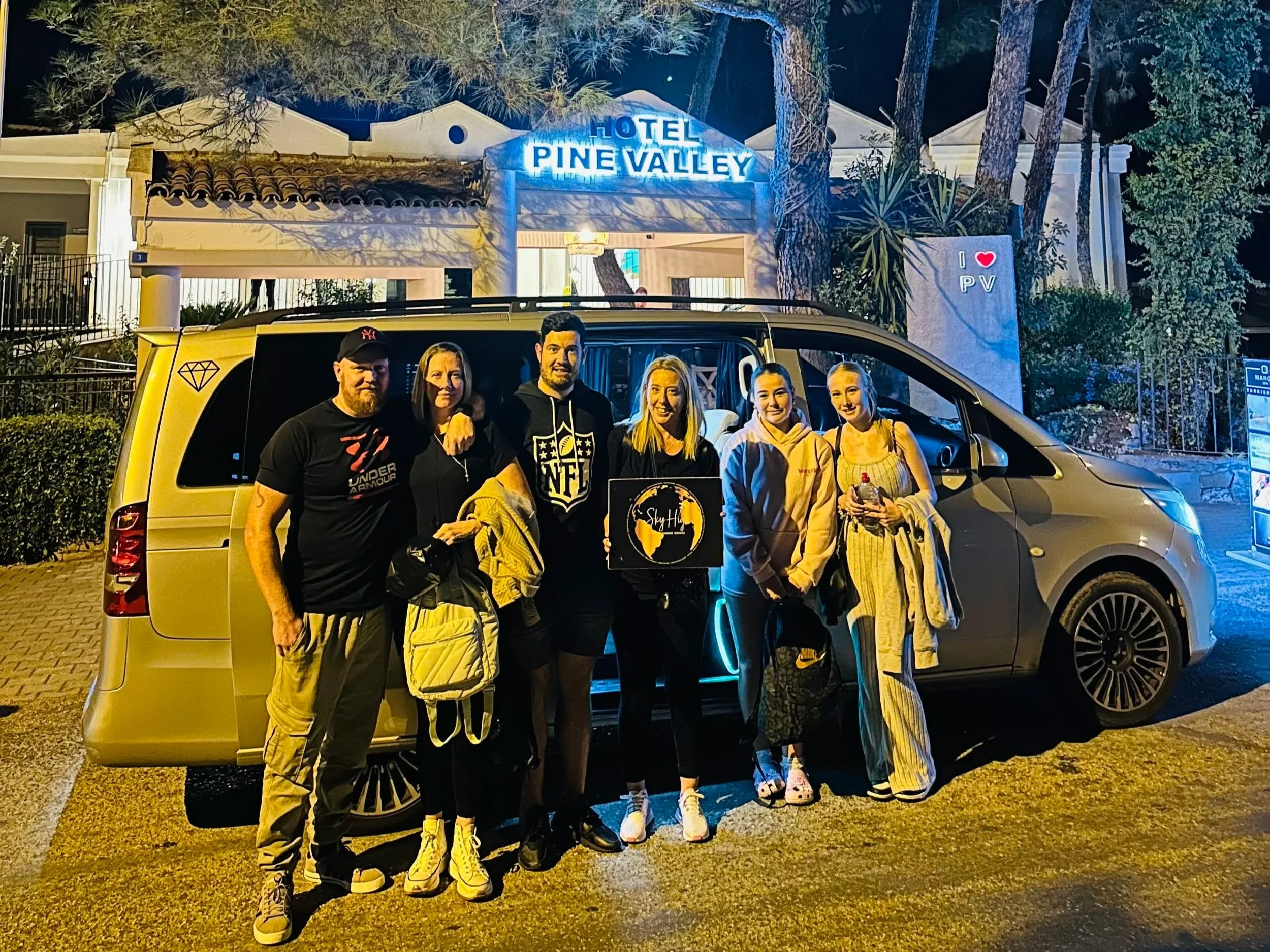 Group of six people standing in front of a silver van parked outside Hotel Pine Valley at night, with a lit blue sign and trees in the background.