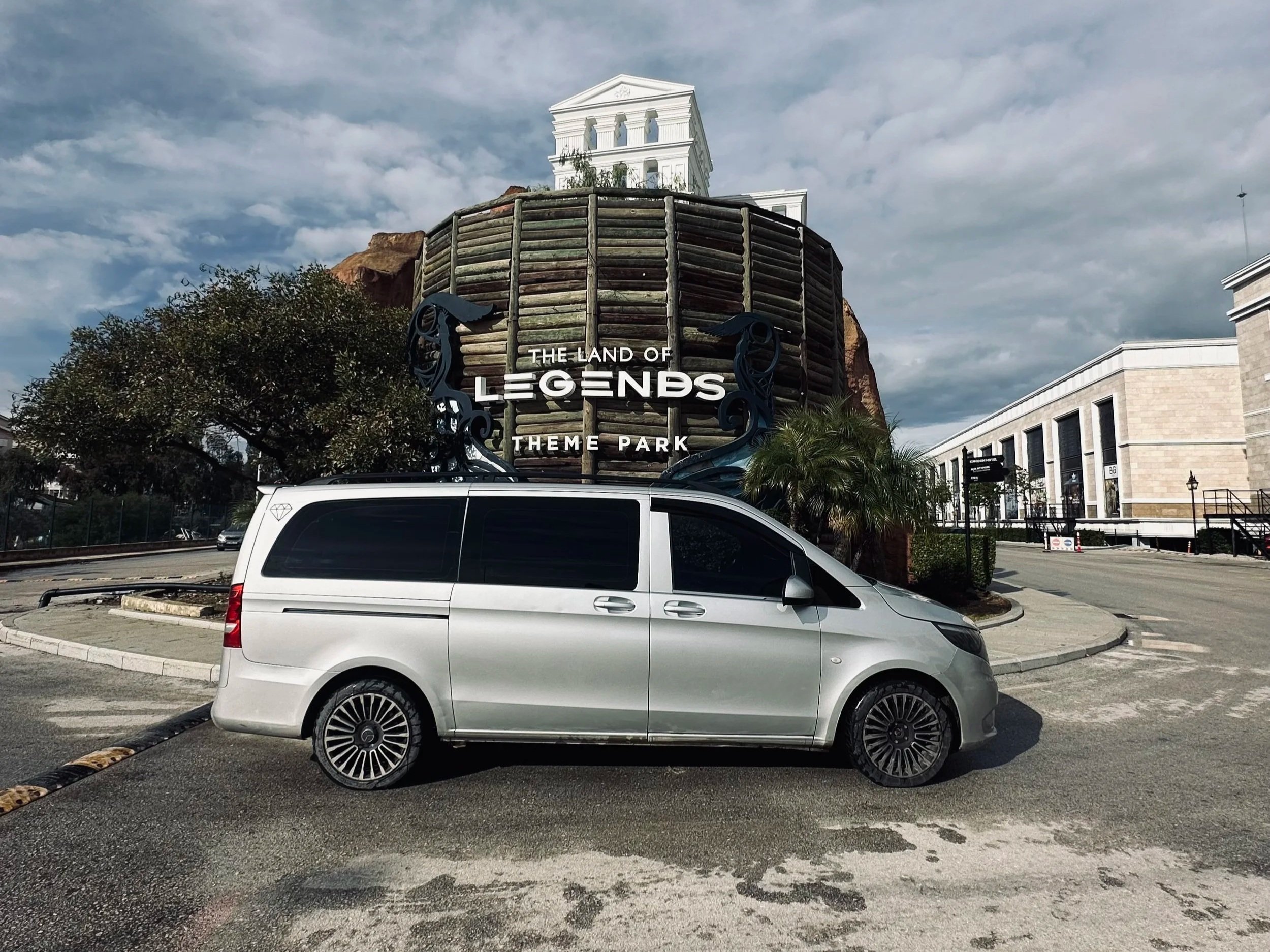 A silver van parked in front of a sign that reads 'The Land of Legends Theme Park'. The sign is attached to a structure resembling a large wooden barrel with a building on top, surrounded by trees and modern buildings under a cloudy sky.
