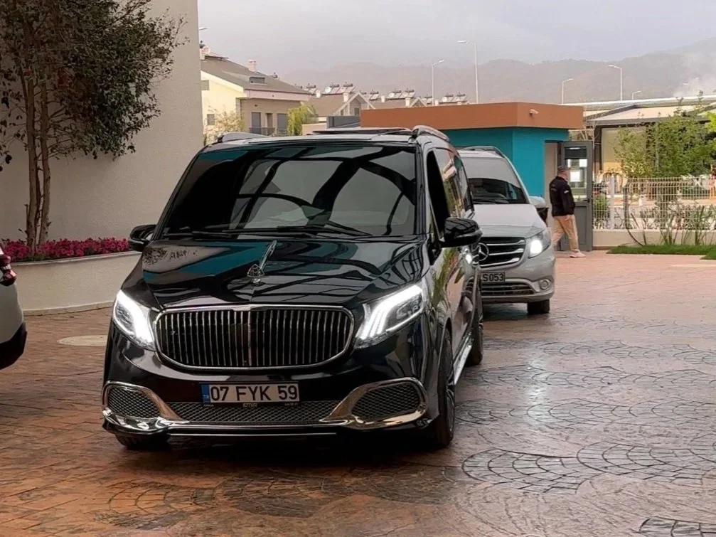 A black luxury van with front license plate 07 FYK 59, parked on a paved area with modern residential buildings and mountains in the background. Reflection of window structures visible on the van's surface.