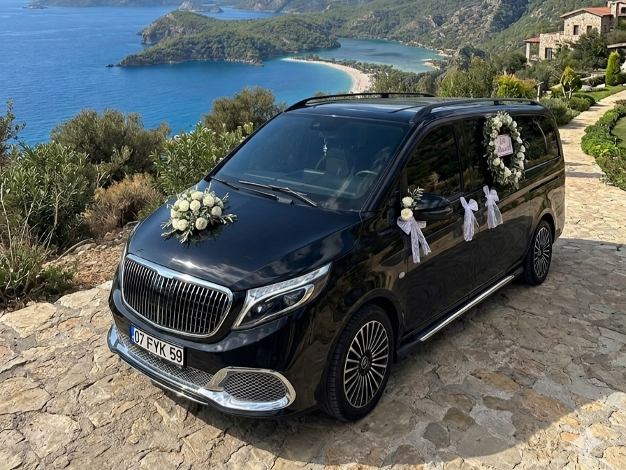 A black hearse decorated with white flowers and ribbons parked on a stone pathway overlooking a scenic coastal view with lush green hills and a blue ocean in the background, suggesting a funeral procession or memorial occasion.