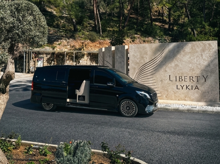 Black luxury van parked in front of a stone wall with the words "Liberty LYKIA" engraved on it, surrounded by landscaped greenery and trees.