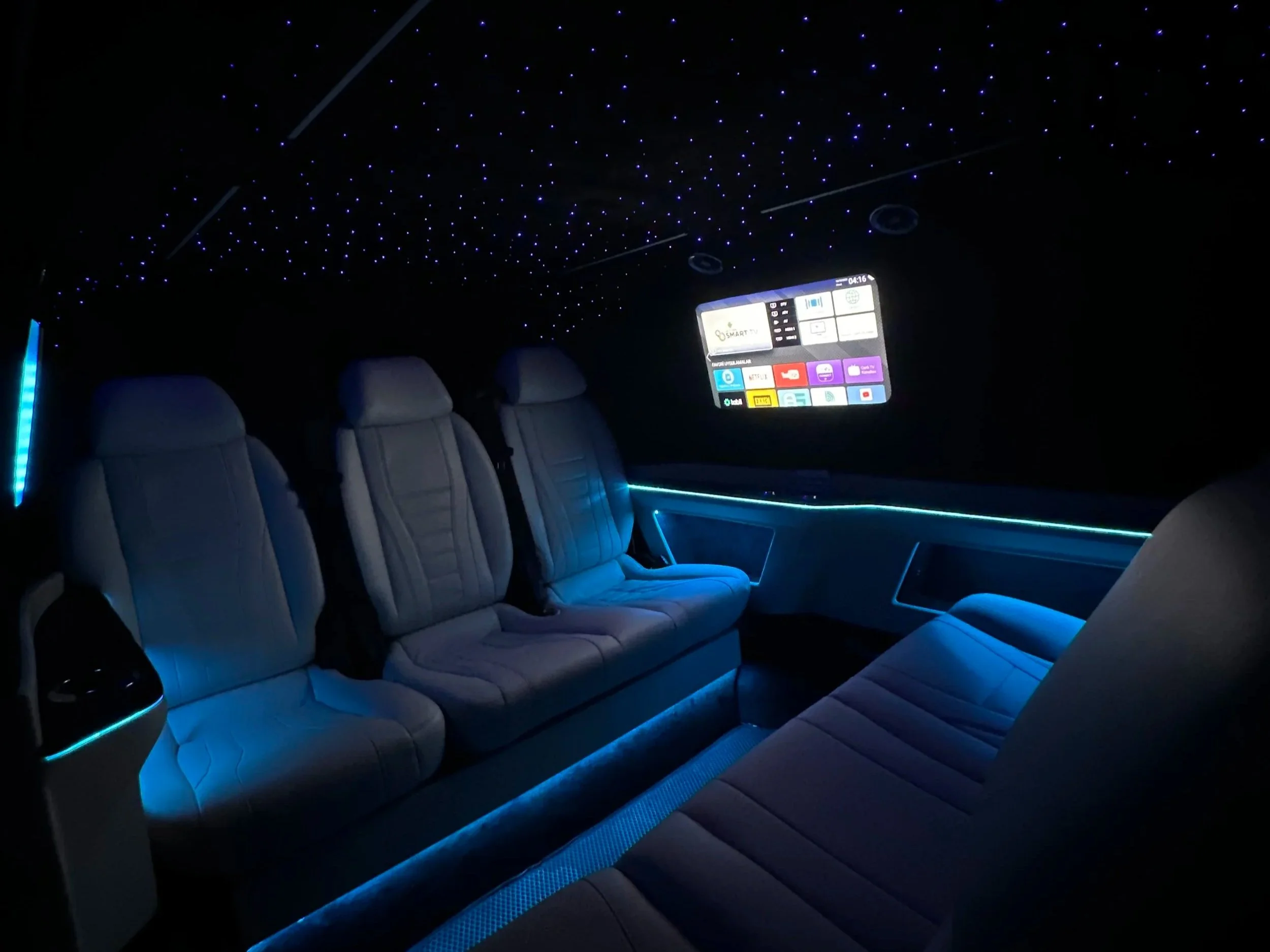 Interior of a luxury vehicle with three gray seats, illuminated ambient lighting, a starry ceiling, and a large digital display screen on the wall.