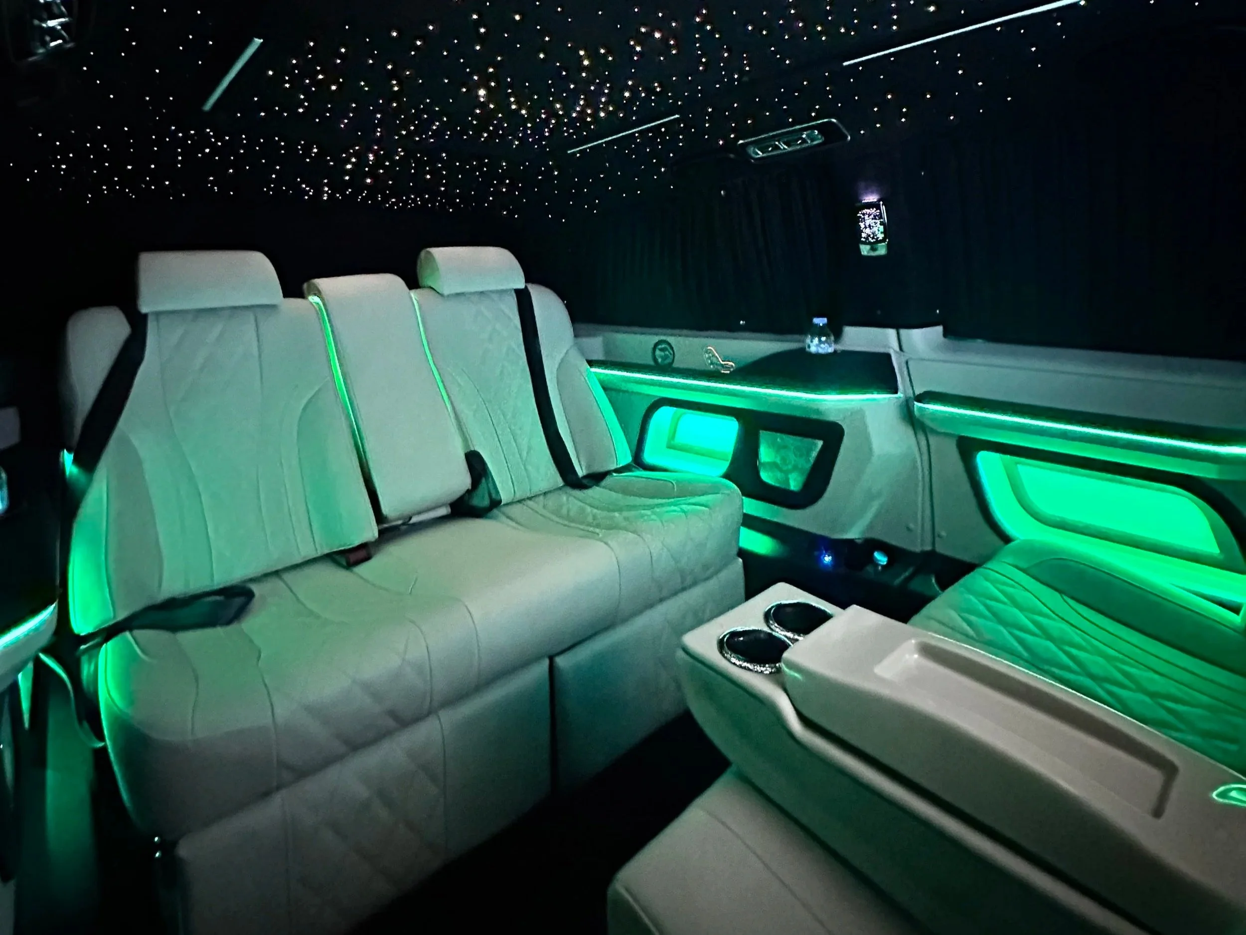 Interior of a luxury limousine with white leather seats, green LED lighting, and a dark ceiling with star-like lights.