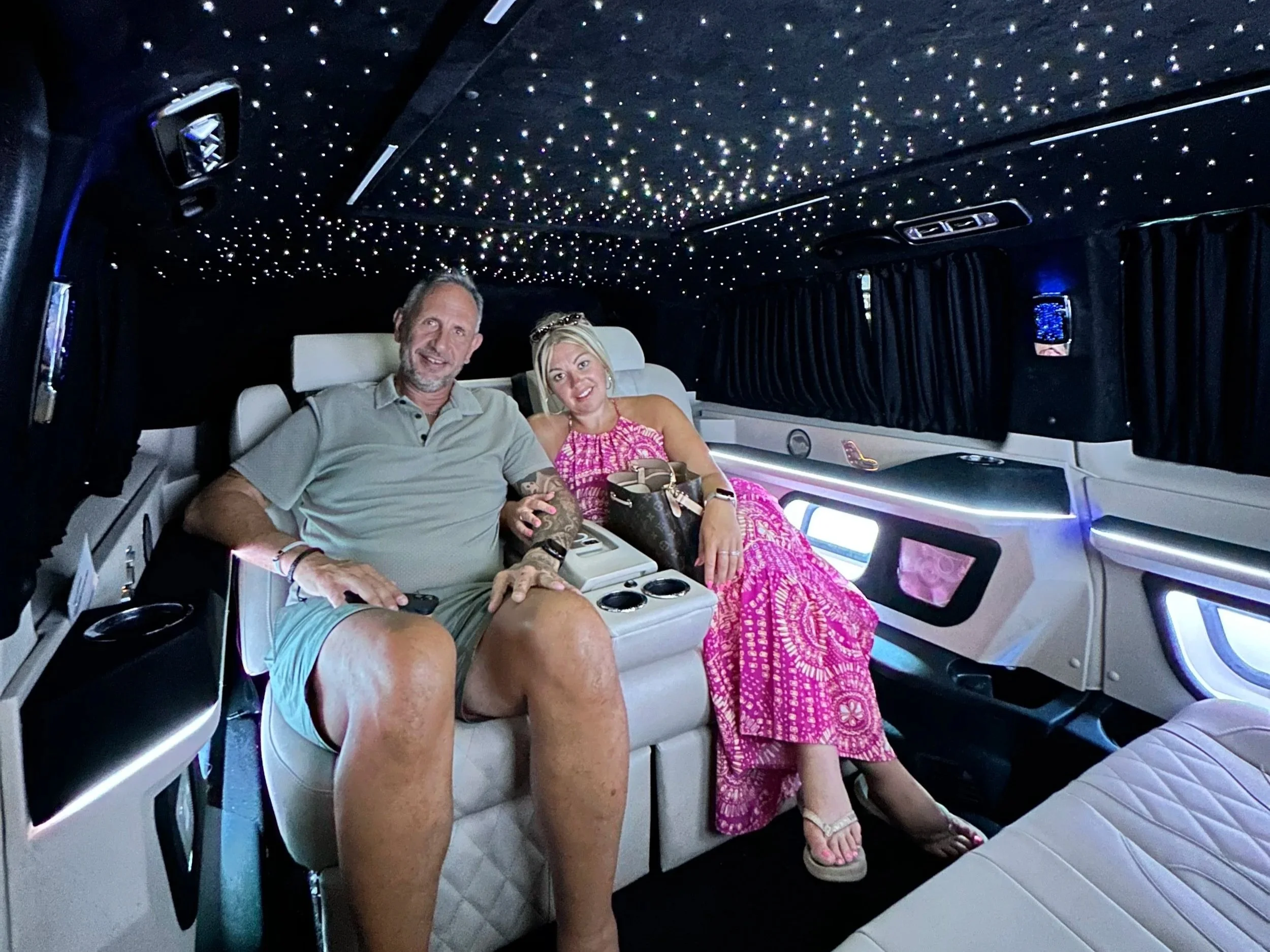 A smiling man and woman sitting inside a limousine with a starry ceiling, black curtains, and luxury interior, enjoying a ride.