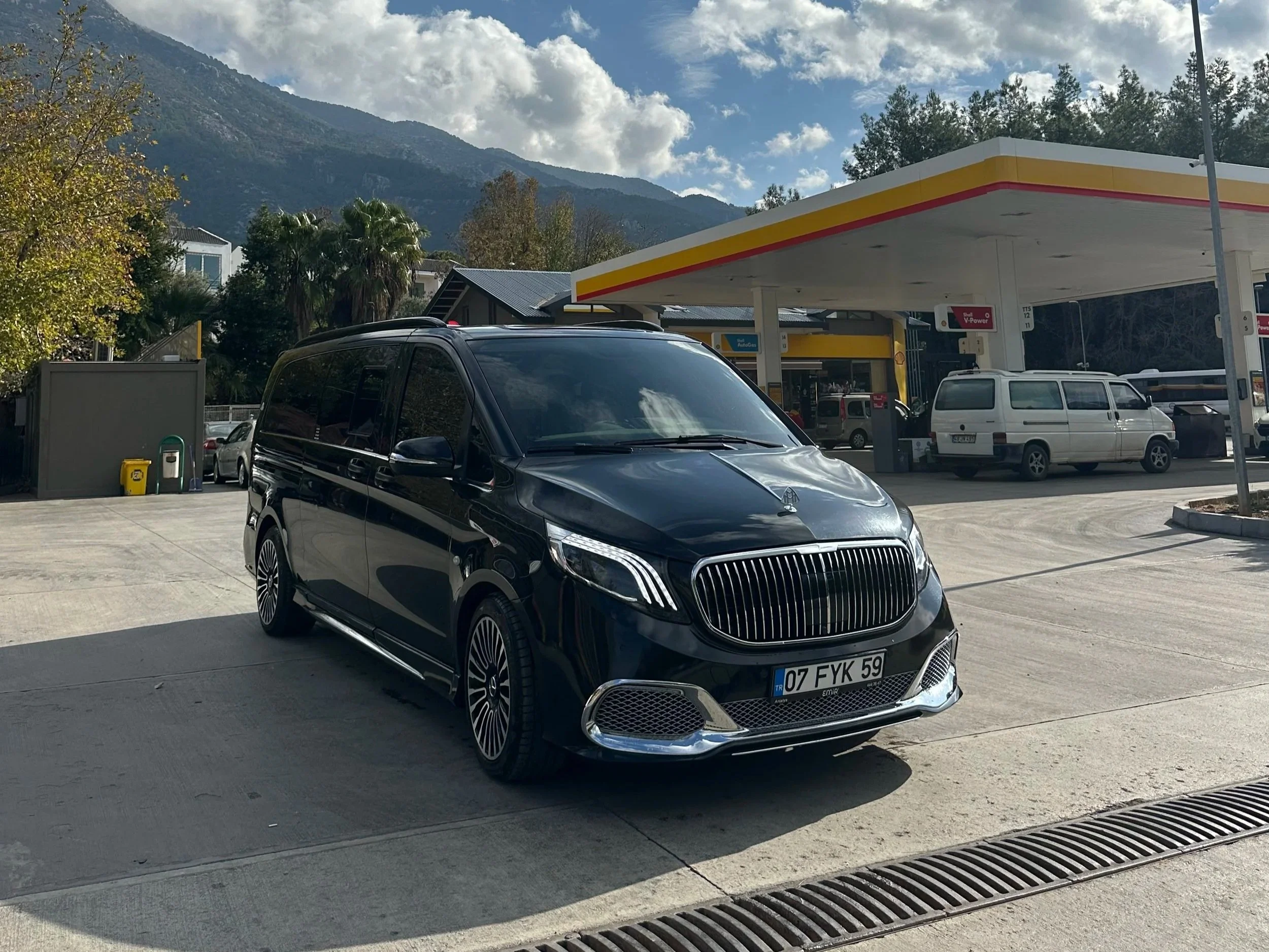 A black luxury van parked at a gas station with a Shell pump in the background, mountains, trees, and a partly cloudy sky overhead.